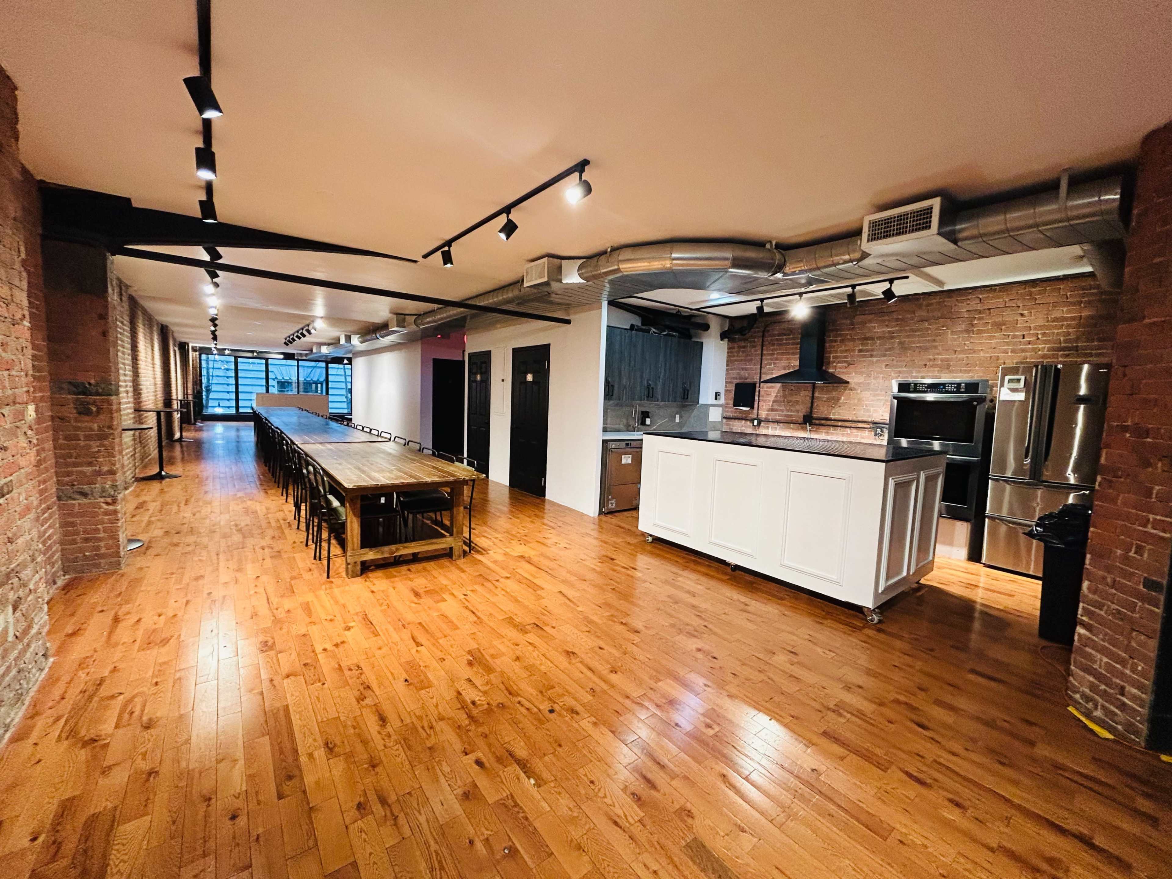 The image shows a spacious, modern kitchen and dining area with exposed brick walls, a long wooden table, and stainless steel appliances.