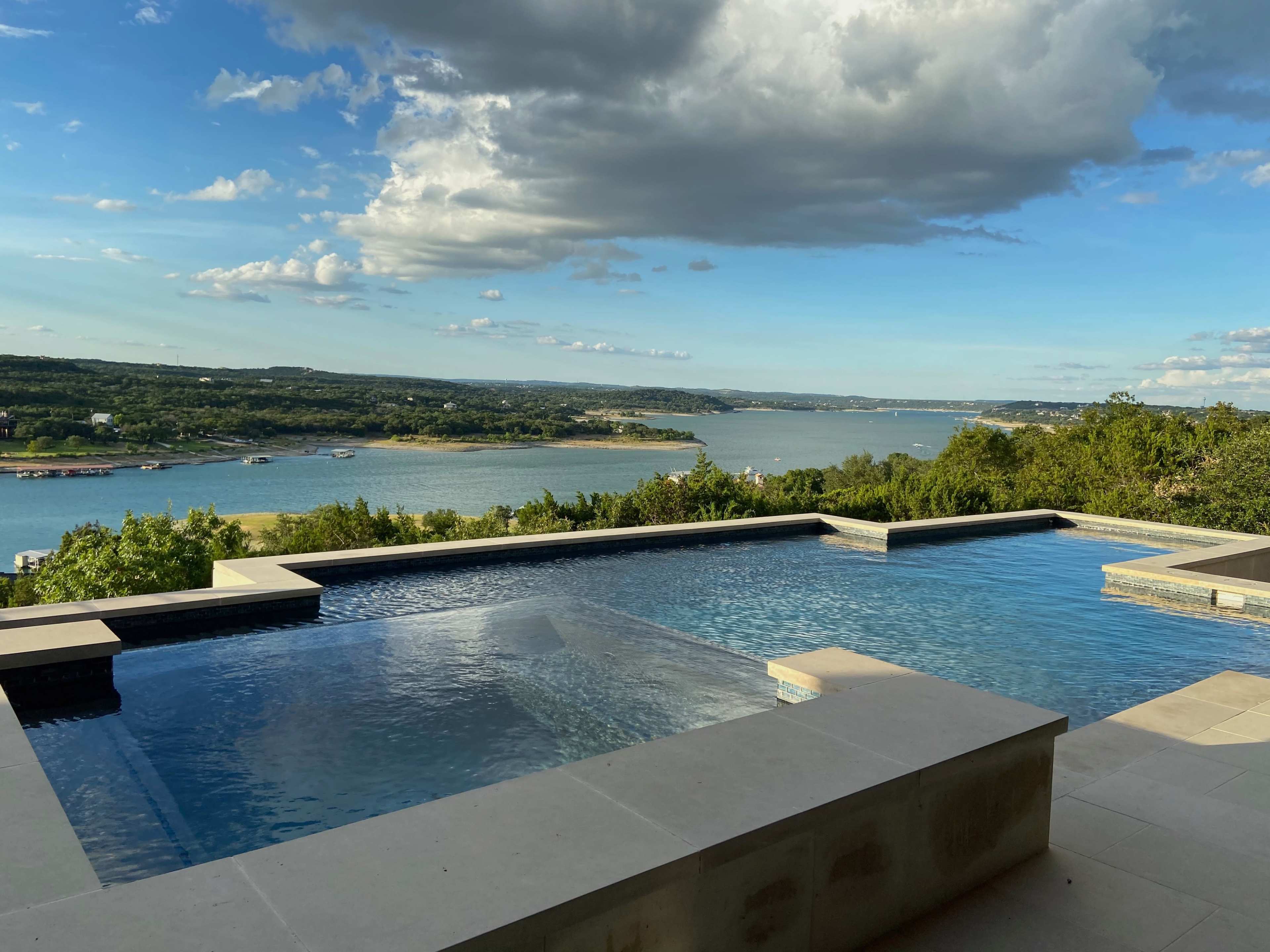The image shows a modern infinity pool overlooking a tranquil lake and green hills under a partly cloudy sky.