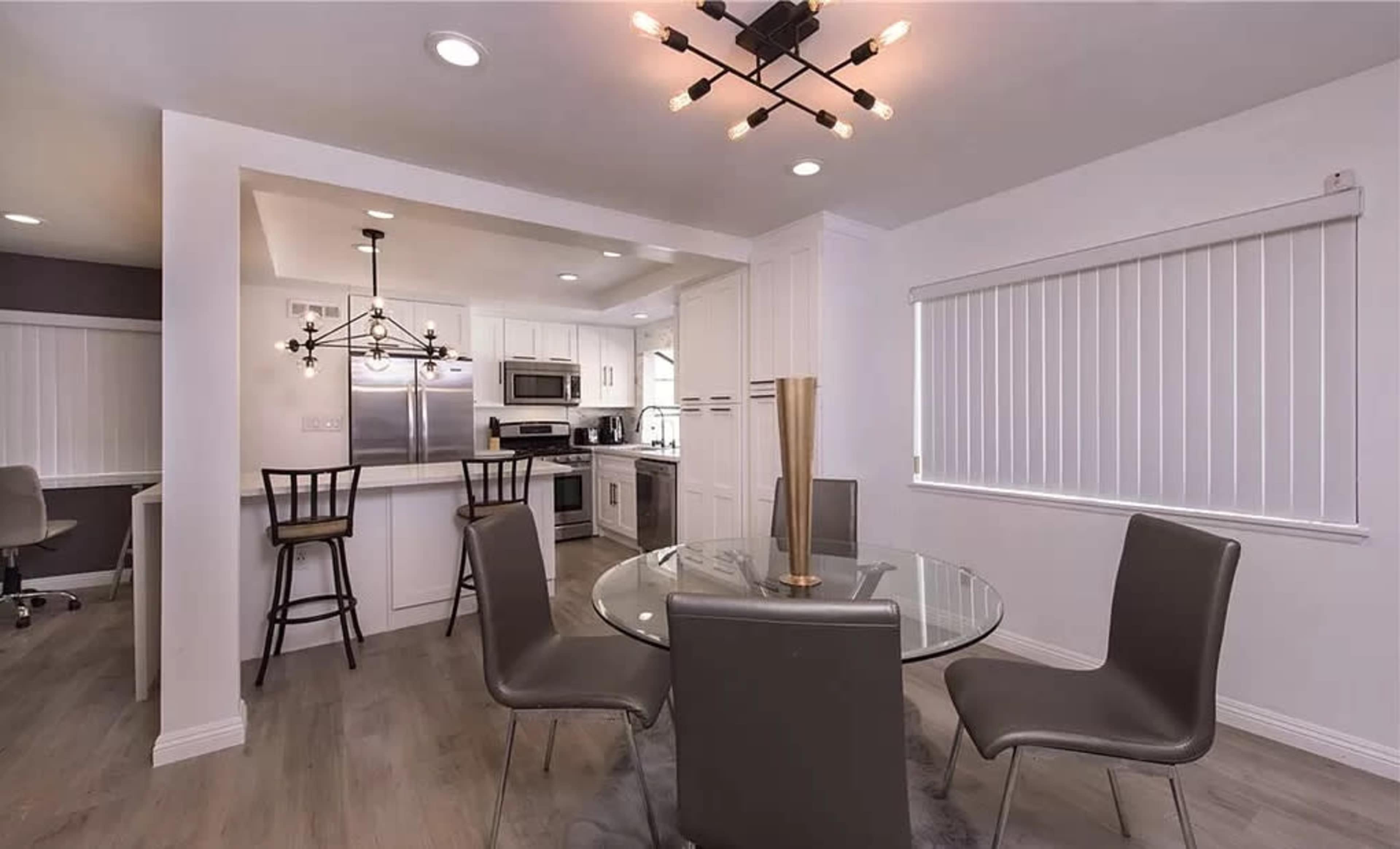 A modern dining area features a round glass table surrounded by four gray chairs, with a kitchen visible in the background.