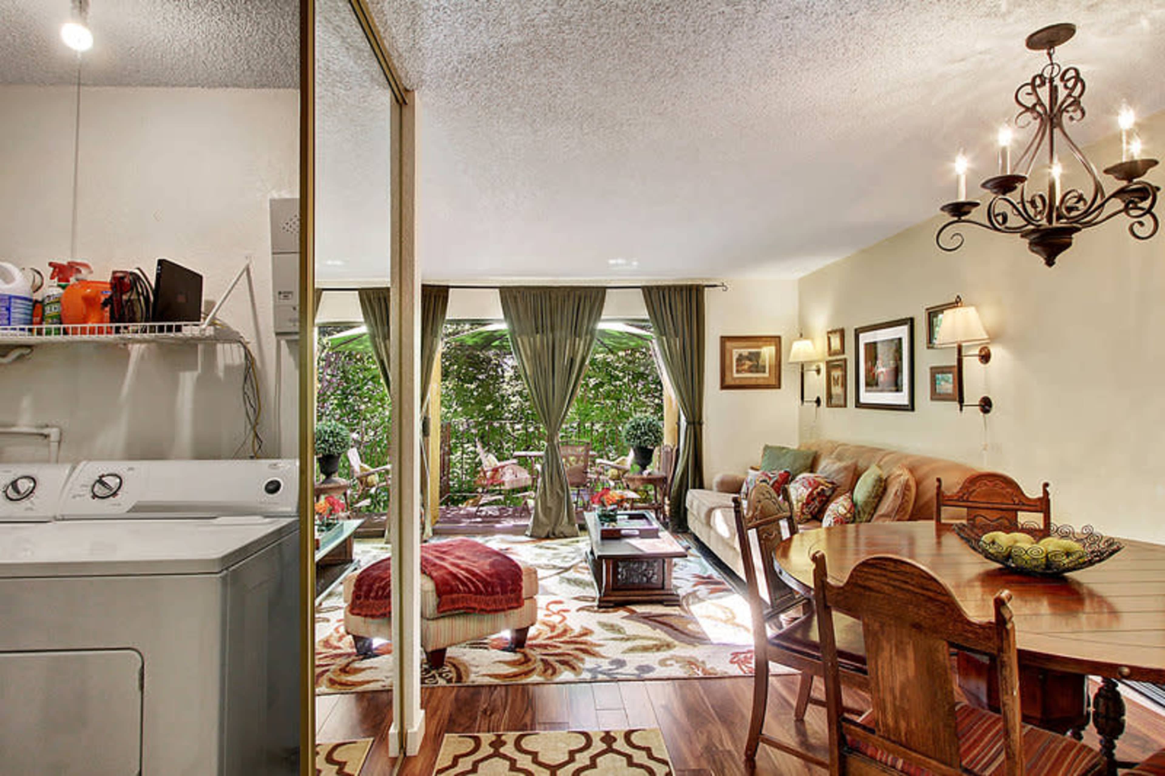 The image shows a living room and dining area with a washer and dryer on one side and a view of a garden through large windows.