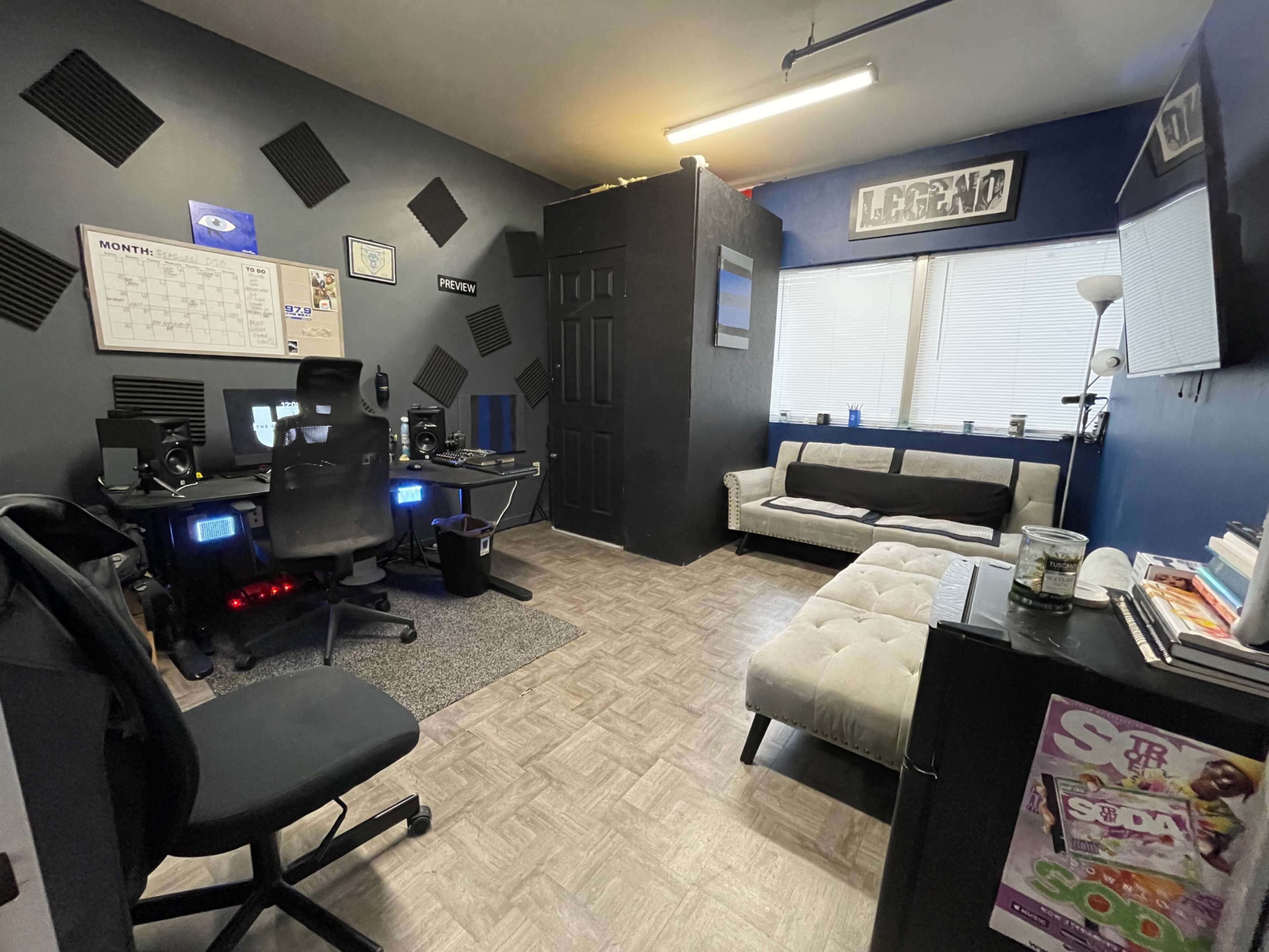 A small, organized room equipped with a desk featuring computer monitors, a seating area with a couch, and sound-proofing panels on the walls.