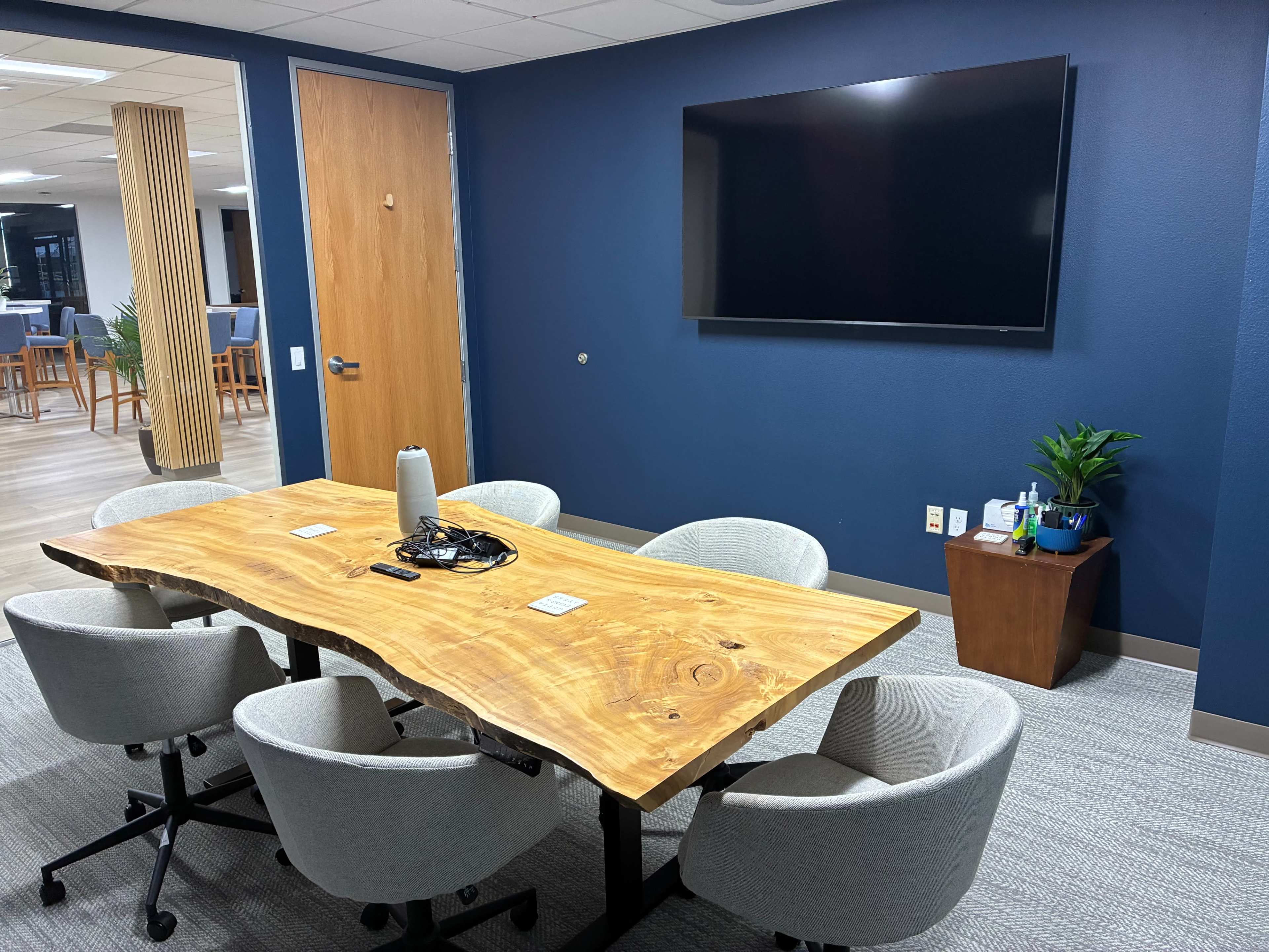 The image shows a modern conference room with a wooden table surrounded by four gray chairs, a wall-mounted television, and a plant in the corner.