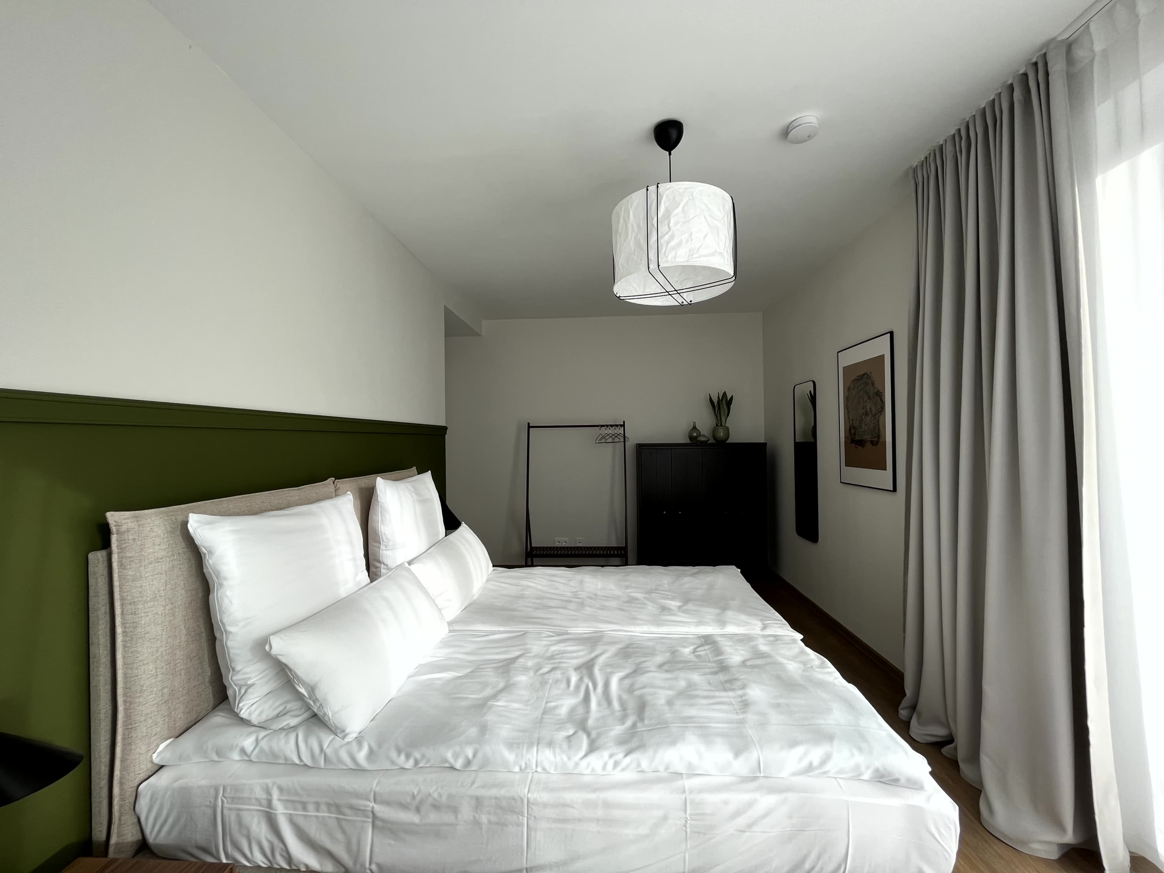 The image shows a neatly arranged bedroom with a double bed, a wardrobe, a hanging clothes rack, and a large window covered by curtains.