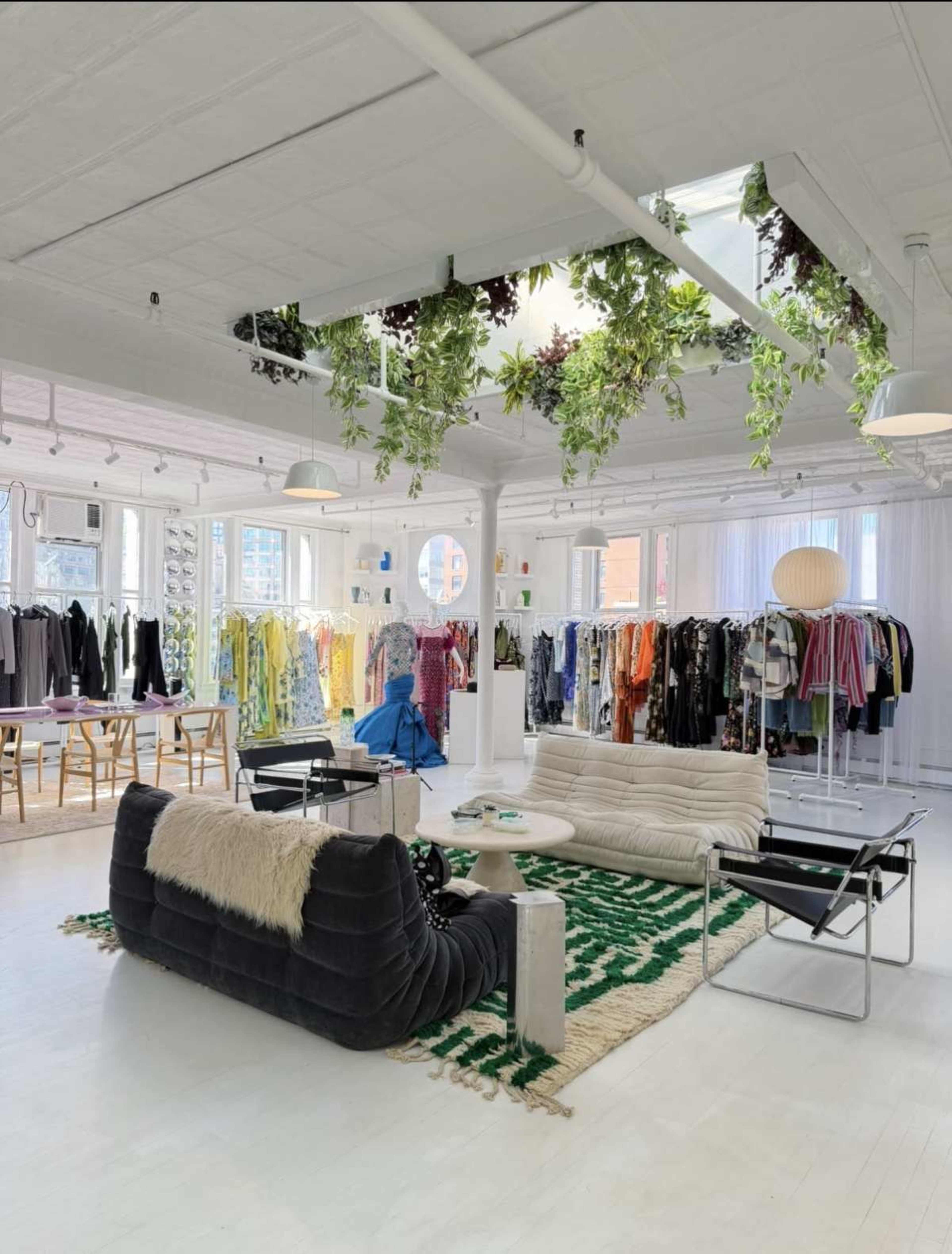 The image shows a modern clothing store with racks of colorful garments, a seating area with contemporary furniture, and greenery hanging from the ceiling.