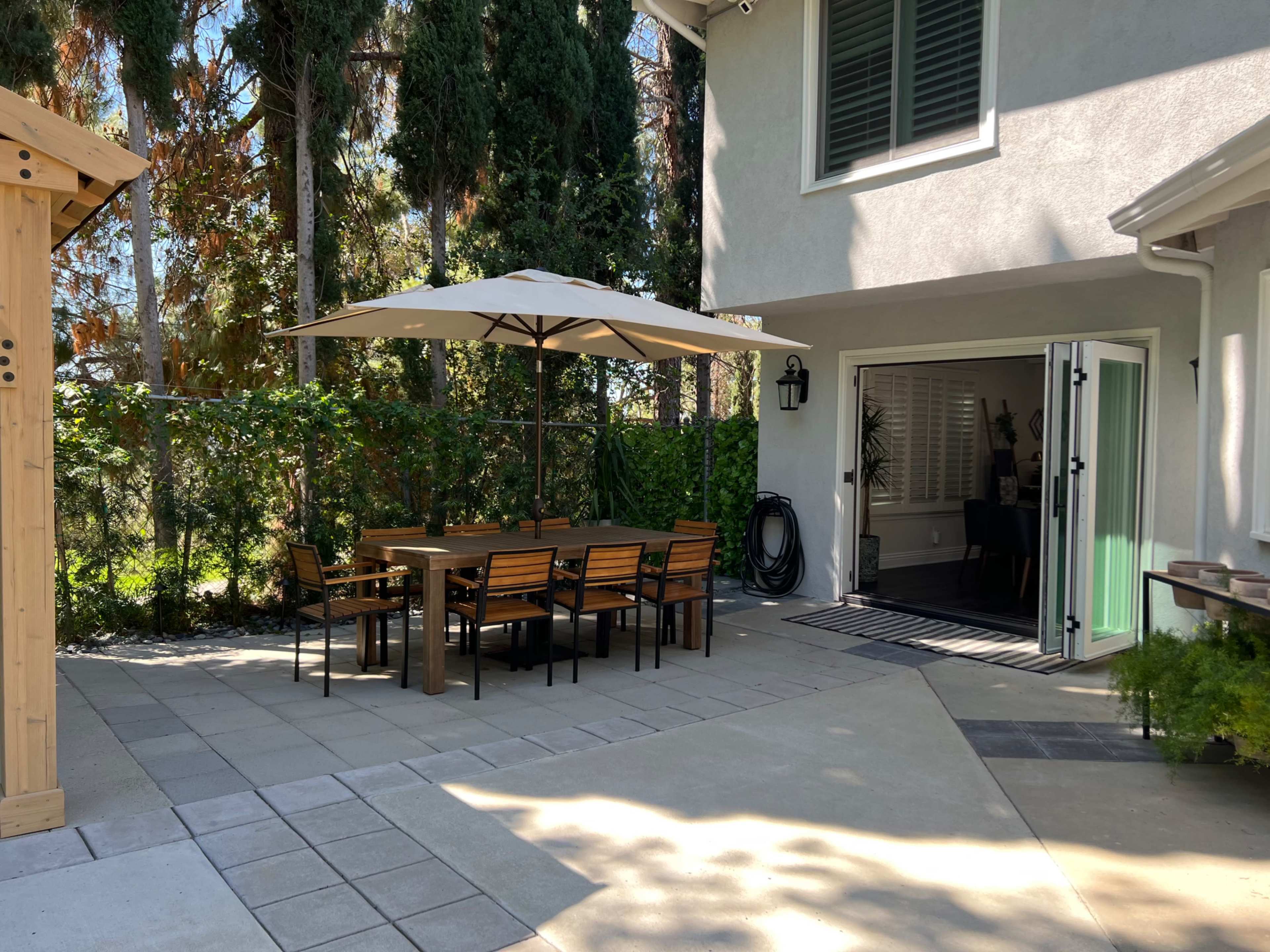 A patio with a wooden dining table and chairs is shaded by a large umbrella, adjacent to a house with open doors leading to an indoor space.
