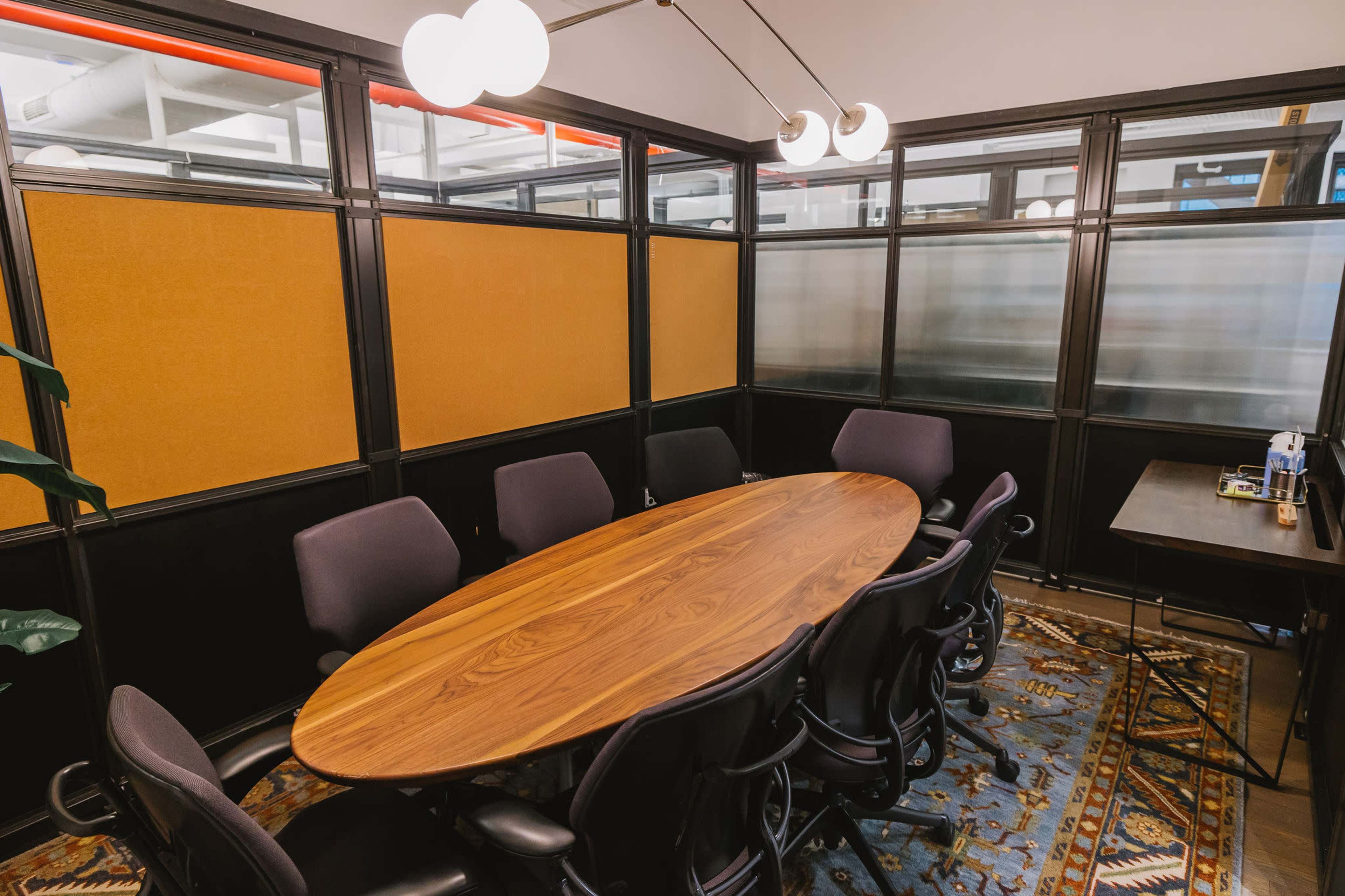 A conference room features a long oval table surrounded by chairs, with glass partitions and modern lighting.
