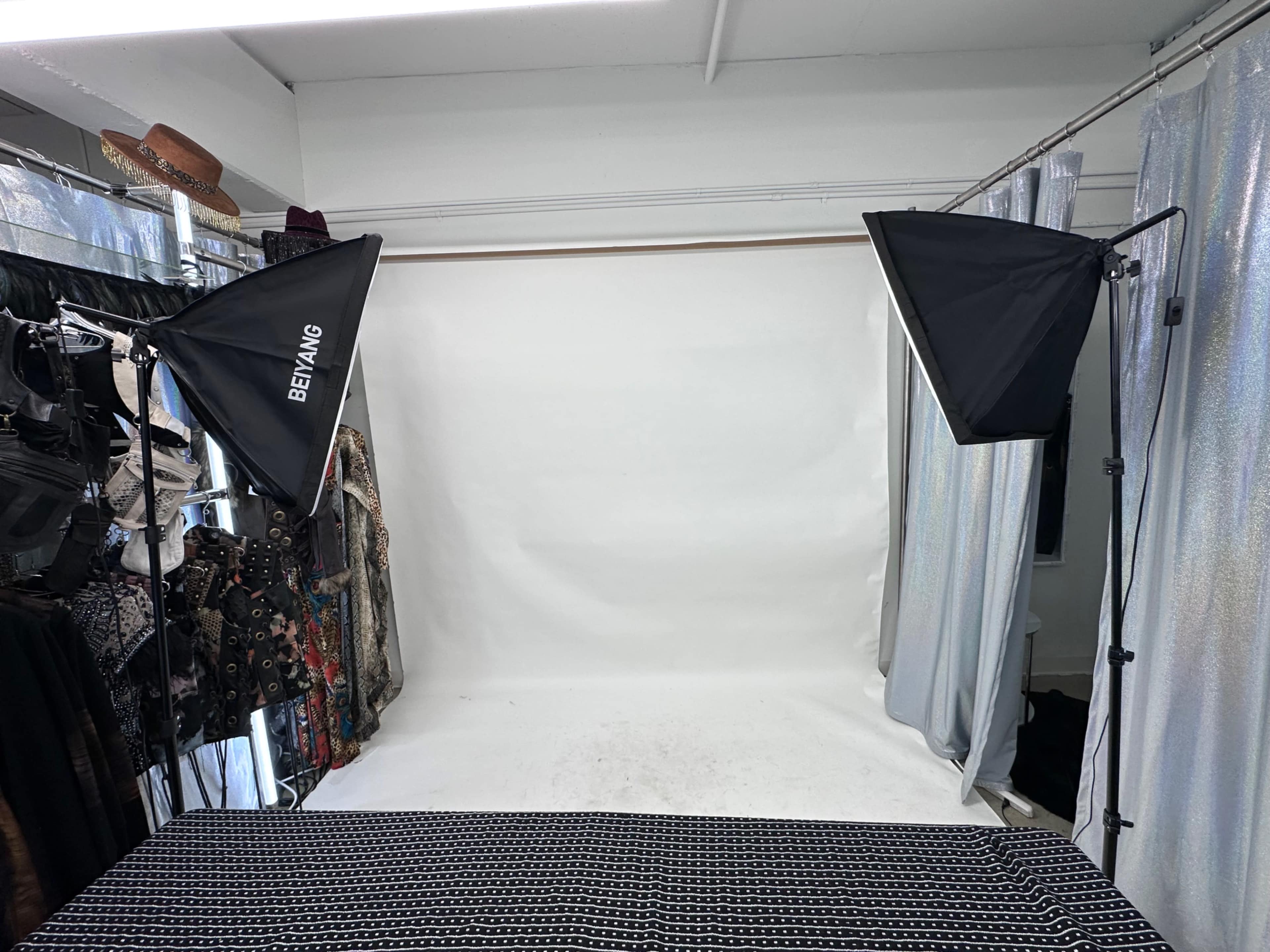 The image shows a photography setup with two softbox lights flanking a backdrop of white paper, surrounded by clothing racks on either side.