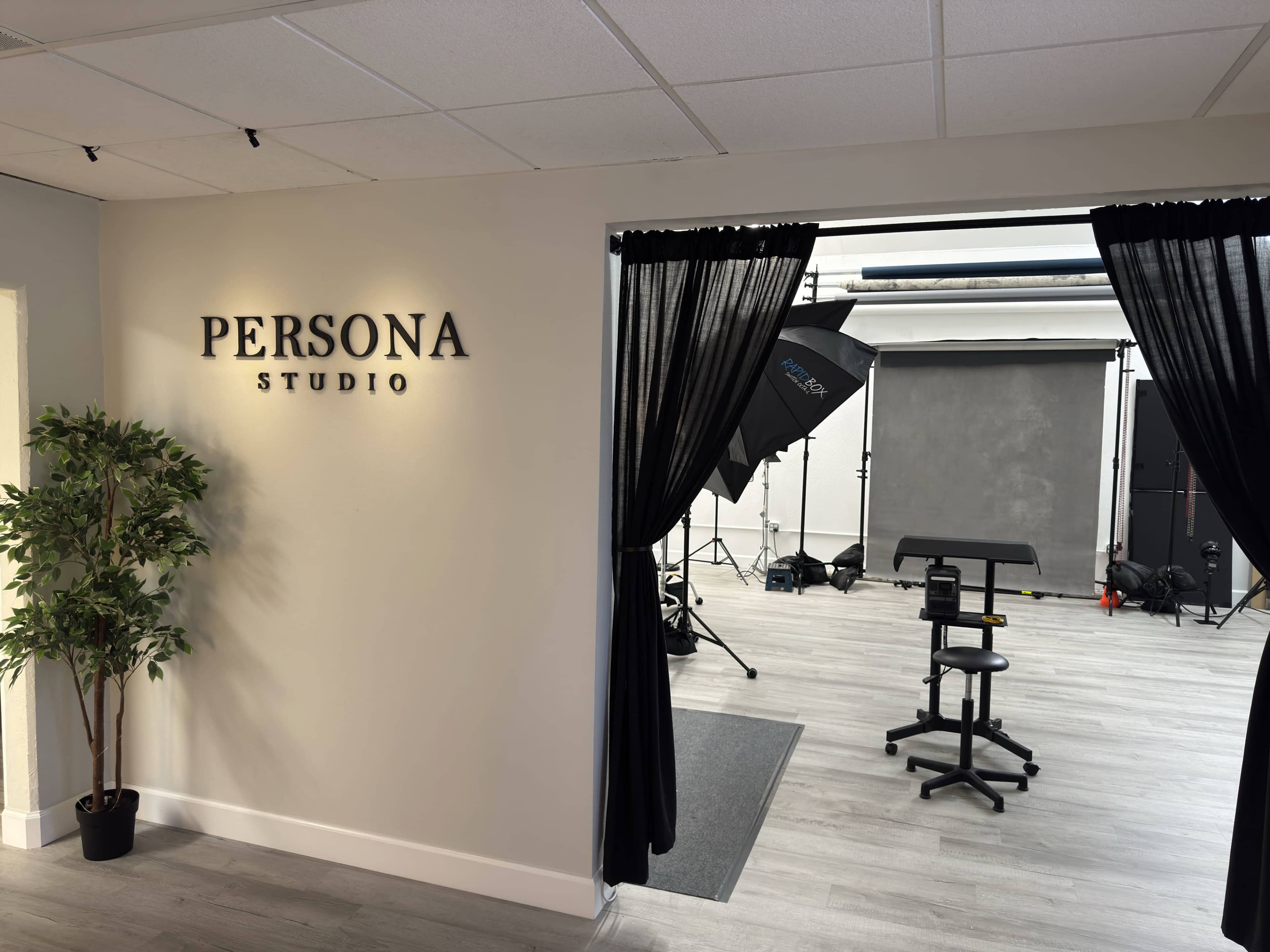 A hallway leads into a studio area with professional lighting equipment, a backdrop, and a chair, framed by black curtains and marked with a sign that says "PERSONA STUDIO."