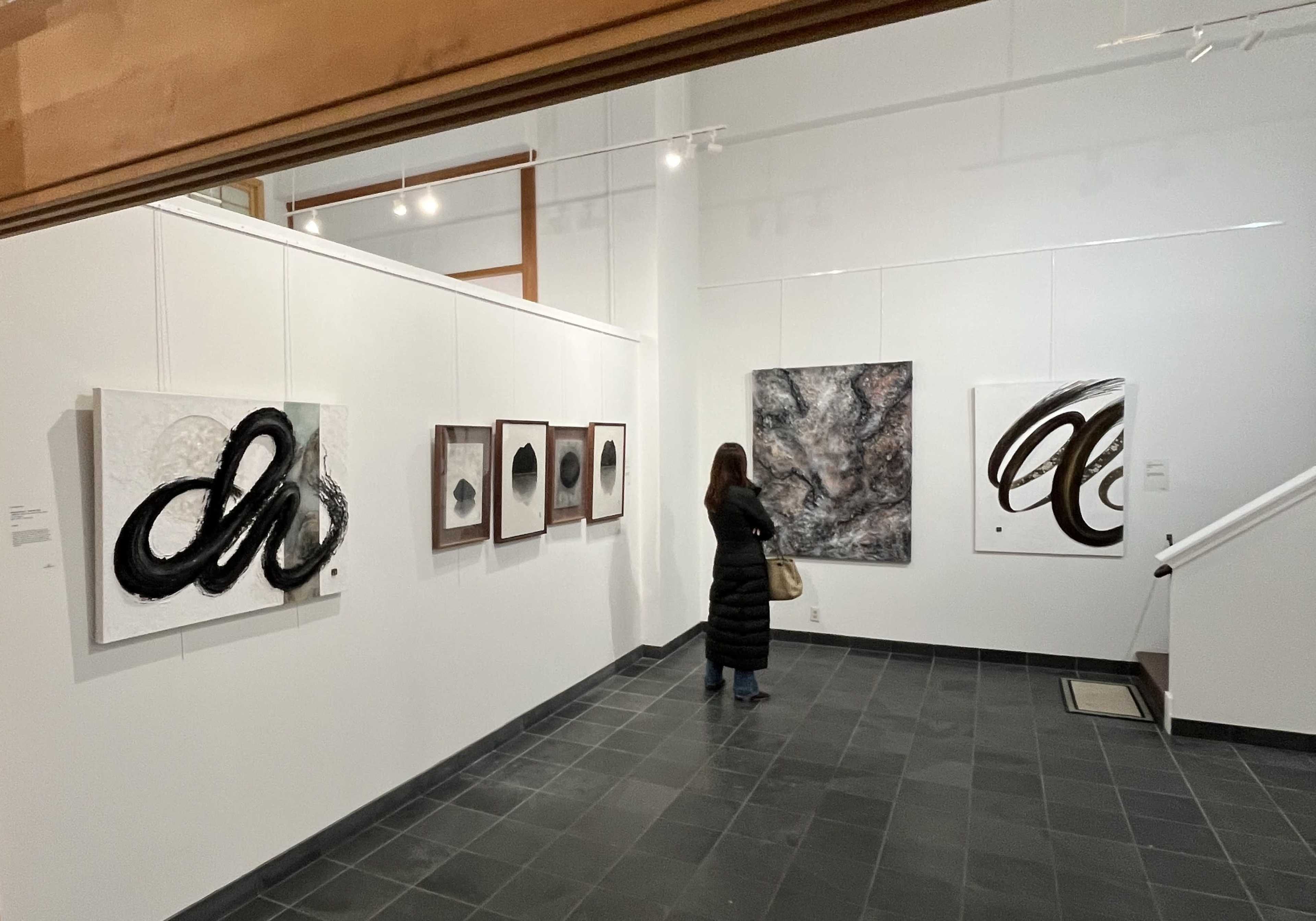 A person stands in an art gallery, observing a large abstract painting on the wall, surrounded by various framed artworks.