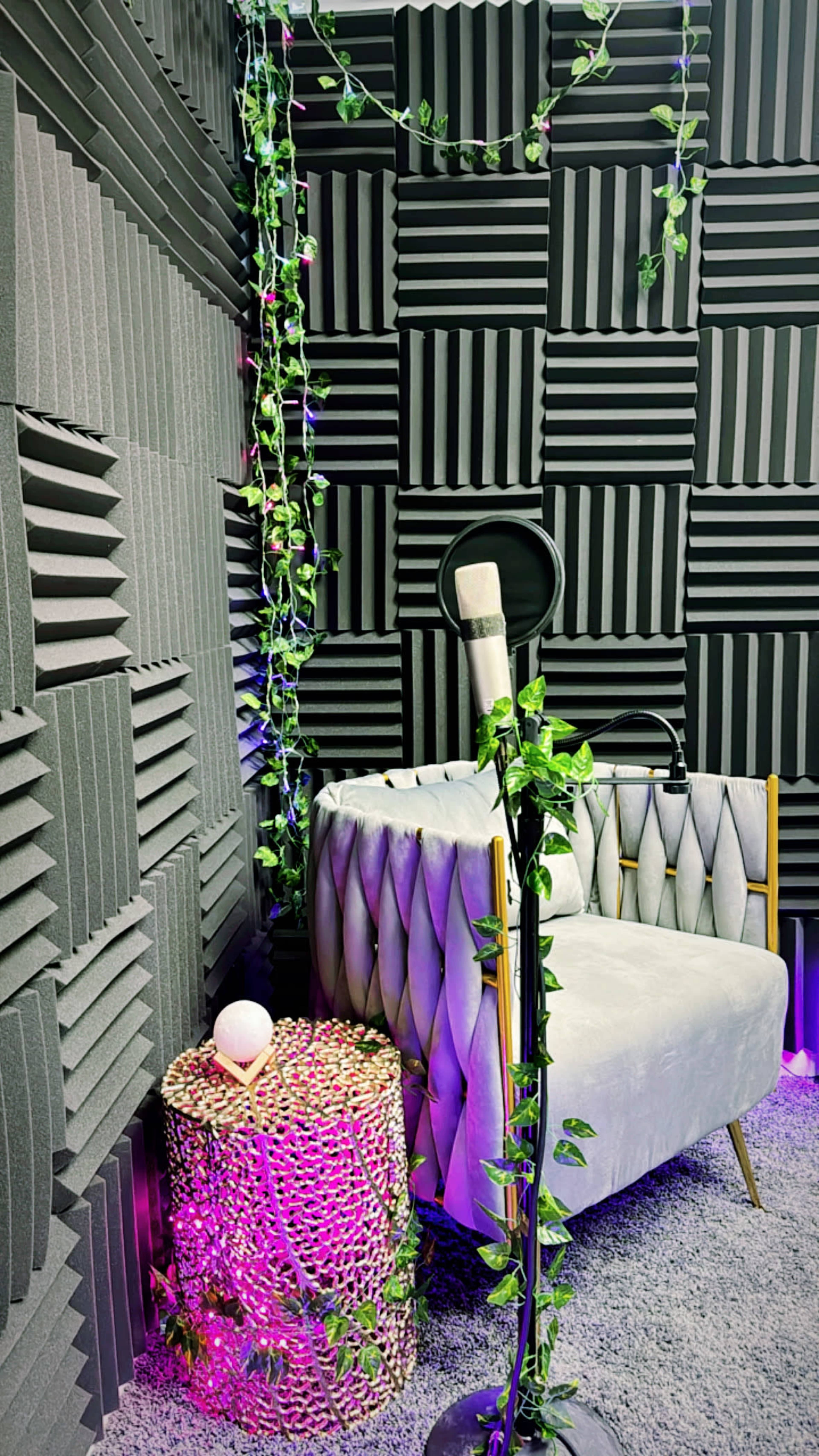 A corner of a soundproofed room with grey acoustic panels, a plush grey chair, a small cylindrical table decorated with lights and greenery, and a microphone on a stand.