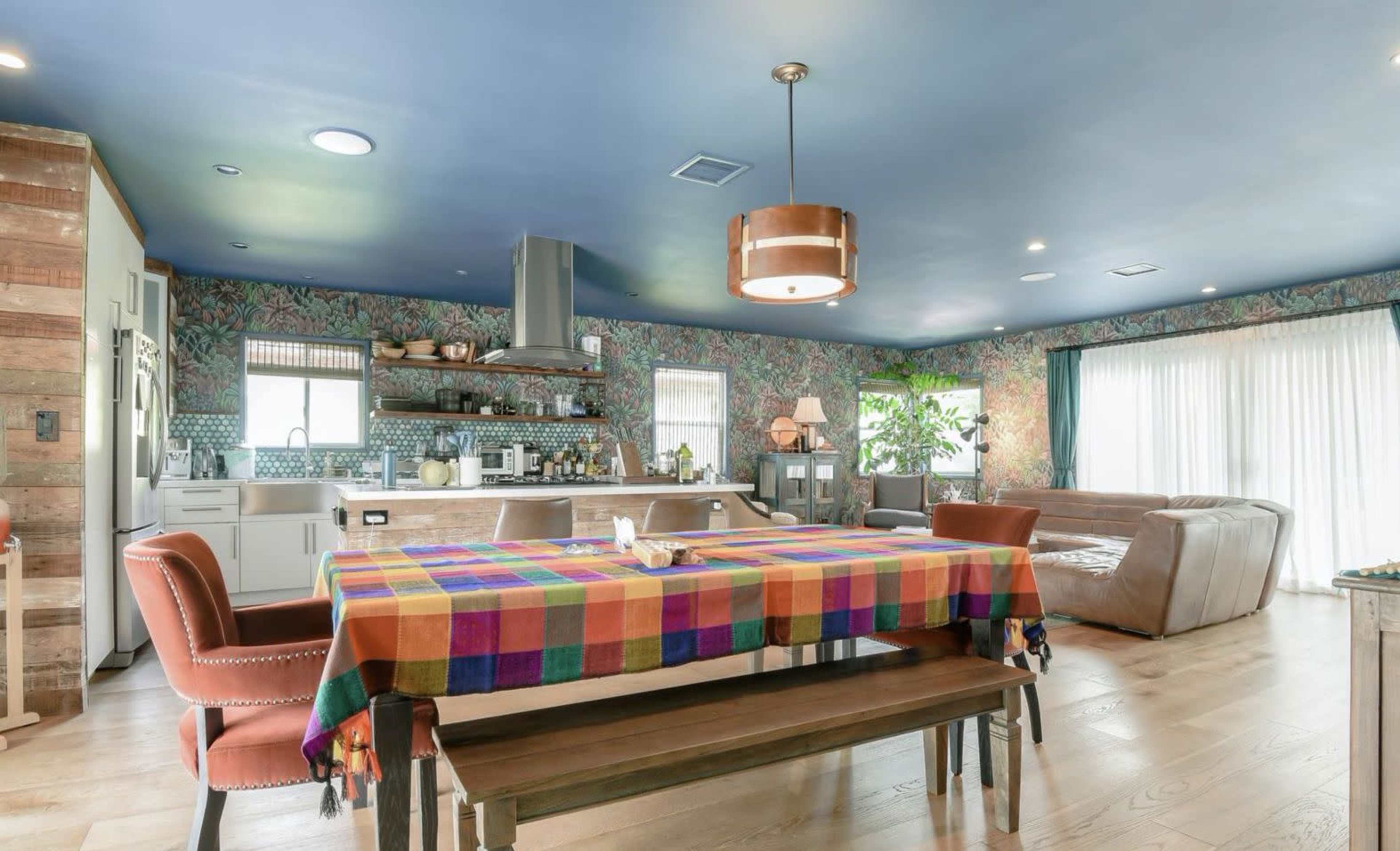 A spacious kitchen and dining area features a colorful tablecloth on a wooden table, surrounded by mixed seating, with floral wallpaper and a large window letting in natural light.