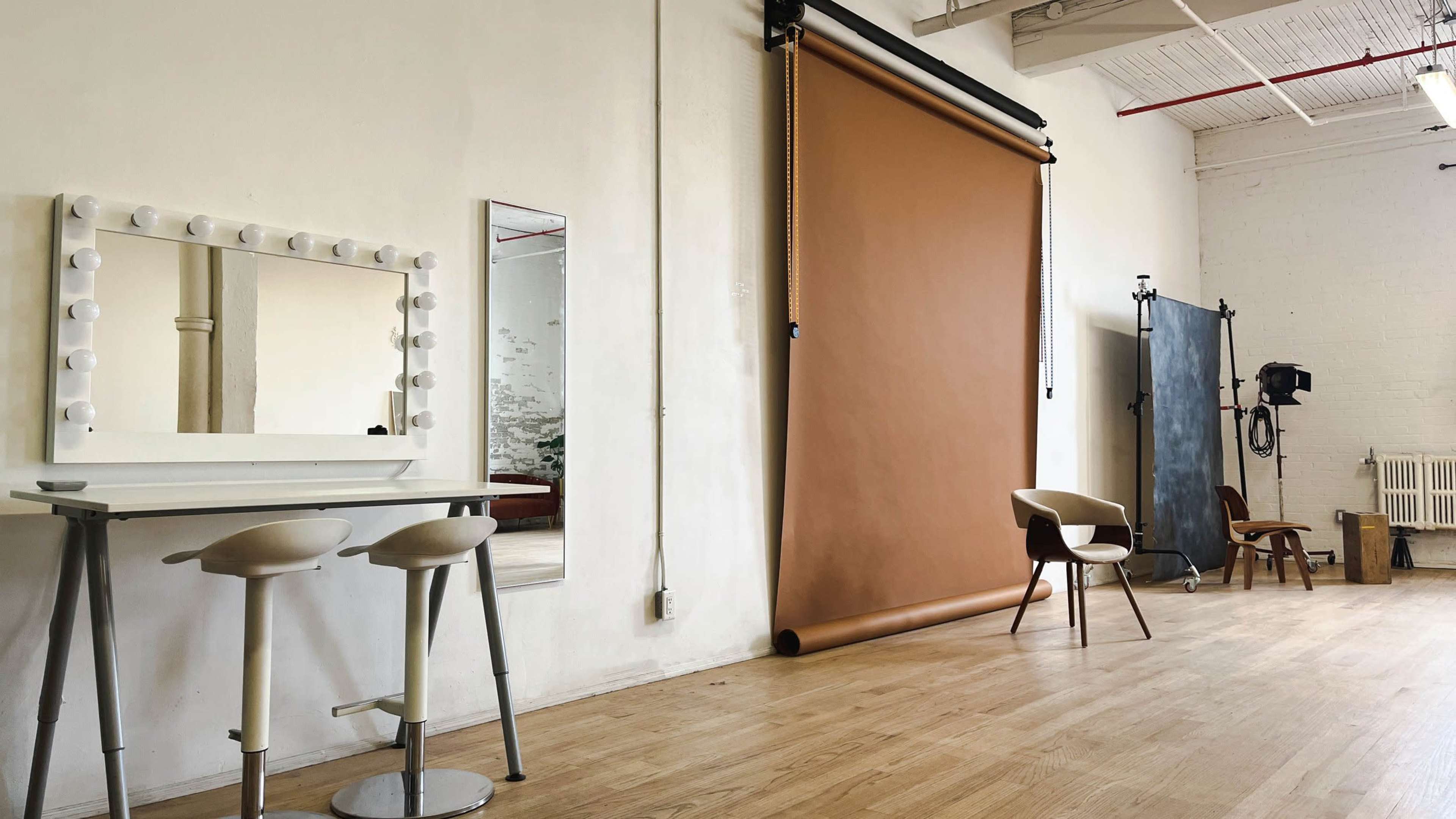 A minimalist studio features a makeup vanity with a mirror, a rolled-up backdrop, and various seating options.
