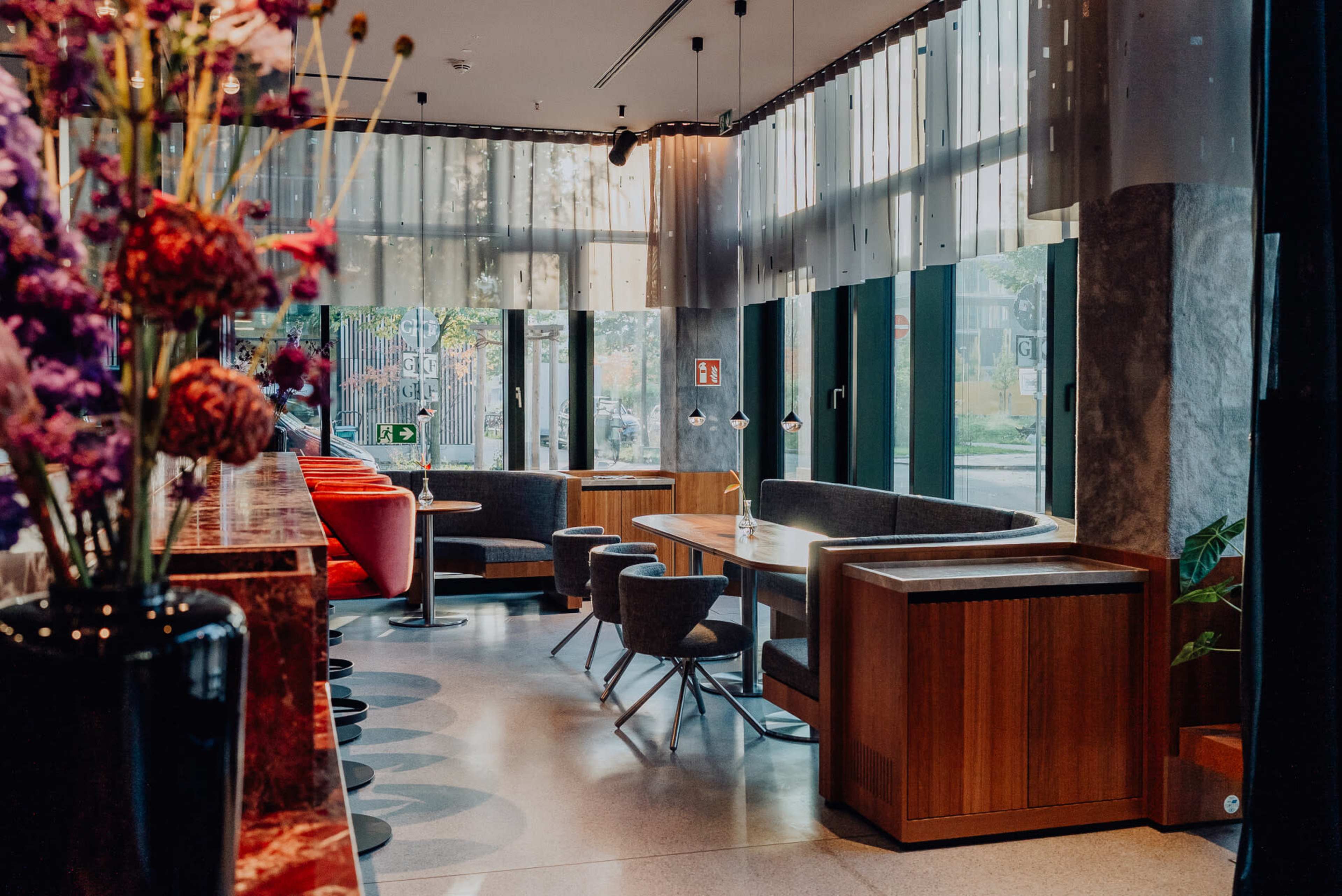 The image shows a modern cafe interior with wooden furniture, large windows, and decorative flowers.