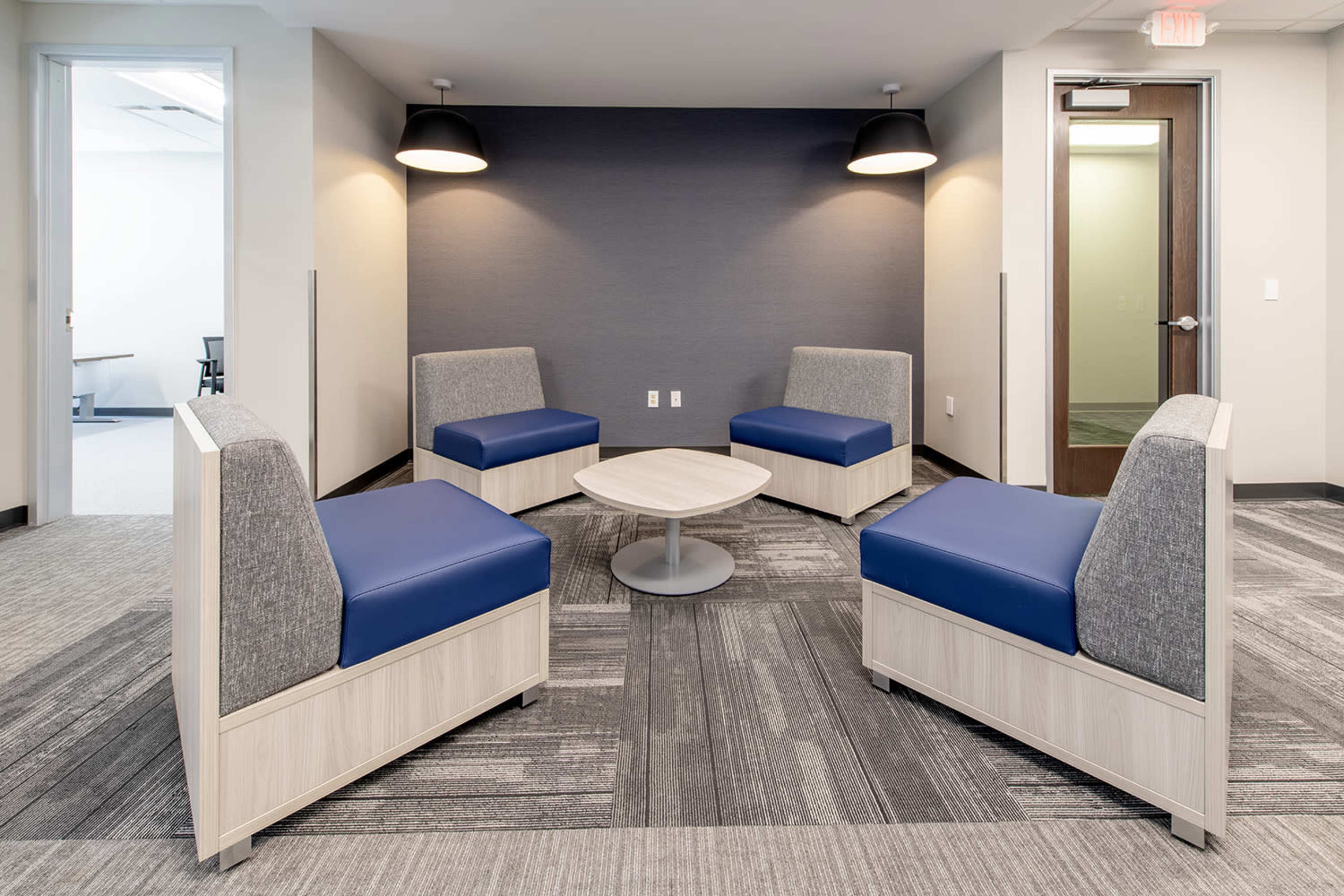 The image shows a modern seating area with four upholstered chairs arranged around a circular table, set against a gray wall and carpeted floor.