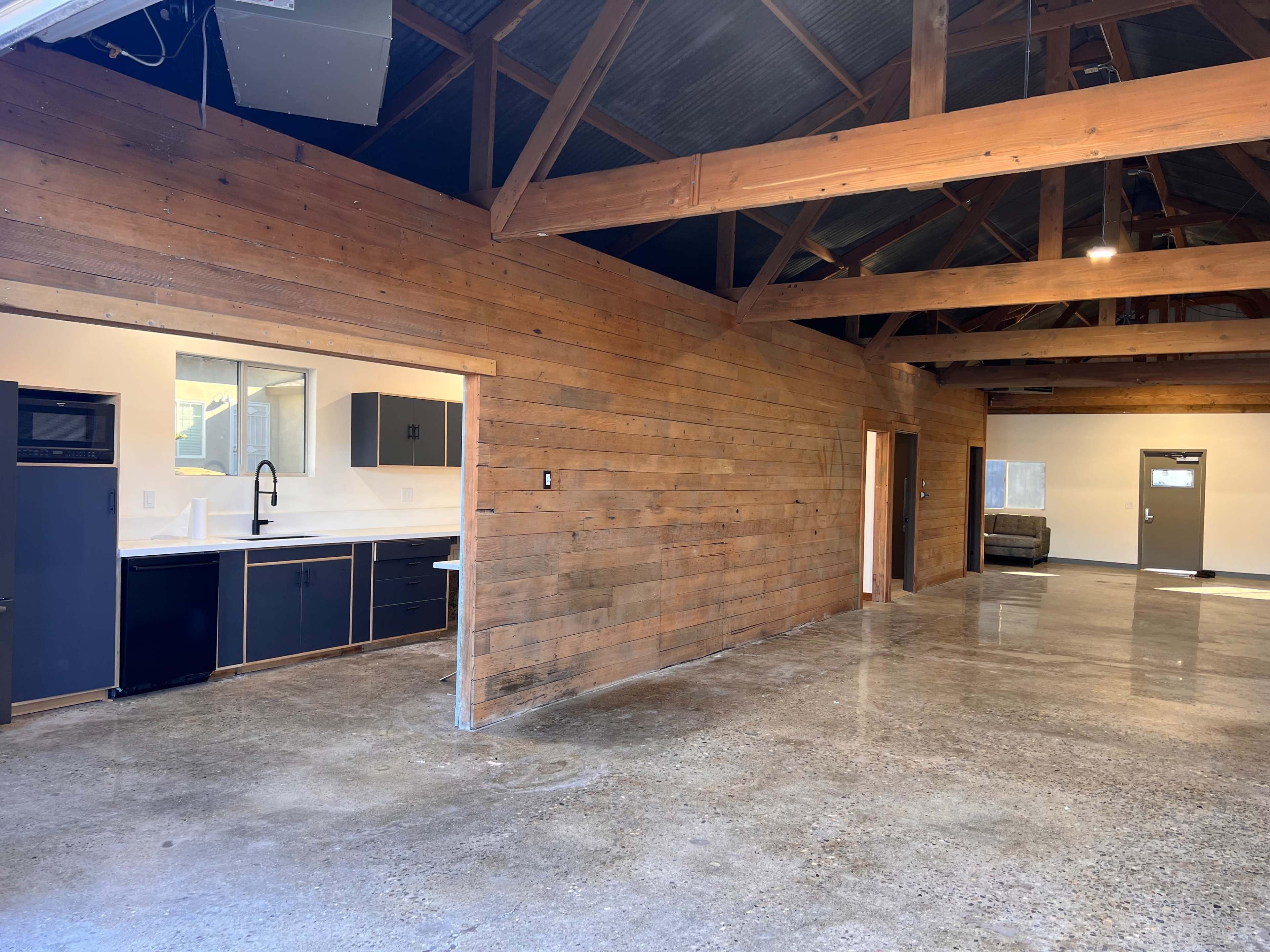 The image shows a spacious room with wooden walls, polished concrete flooring, and a kitchen area featuring dark cabinetry and modern fixtures.