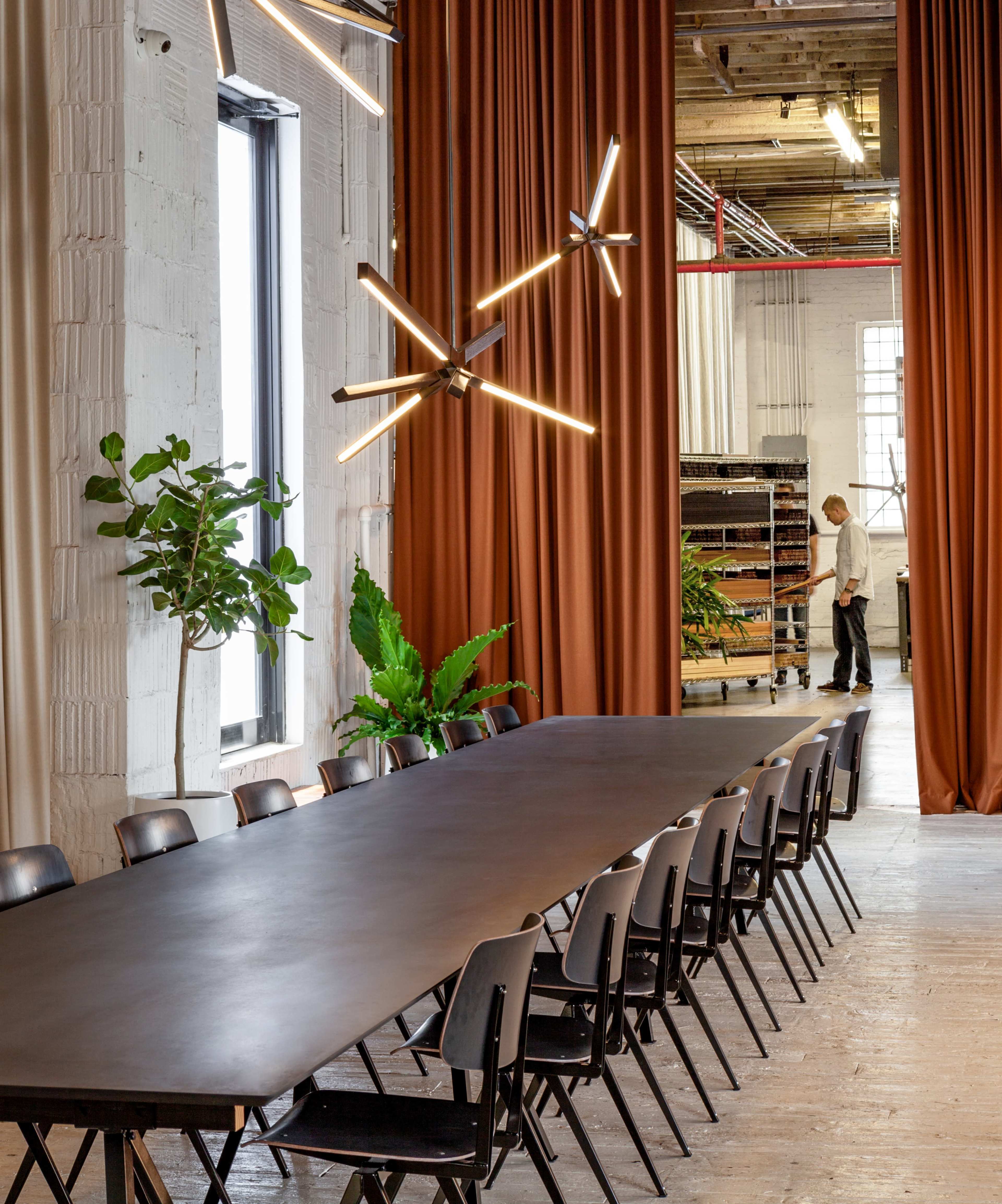 A long black table is set in a well-lit room with curtains, plants, and a person standing nearby.