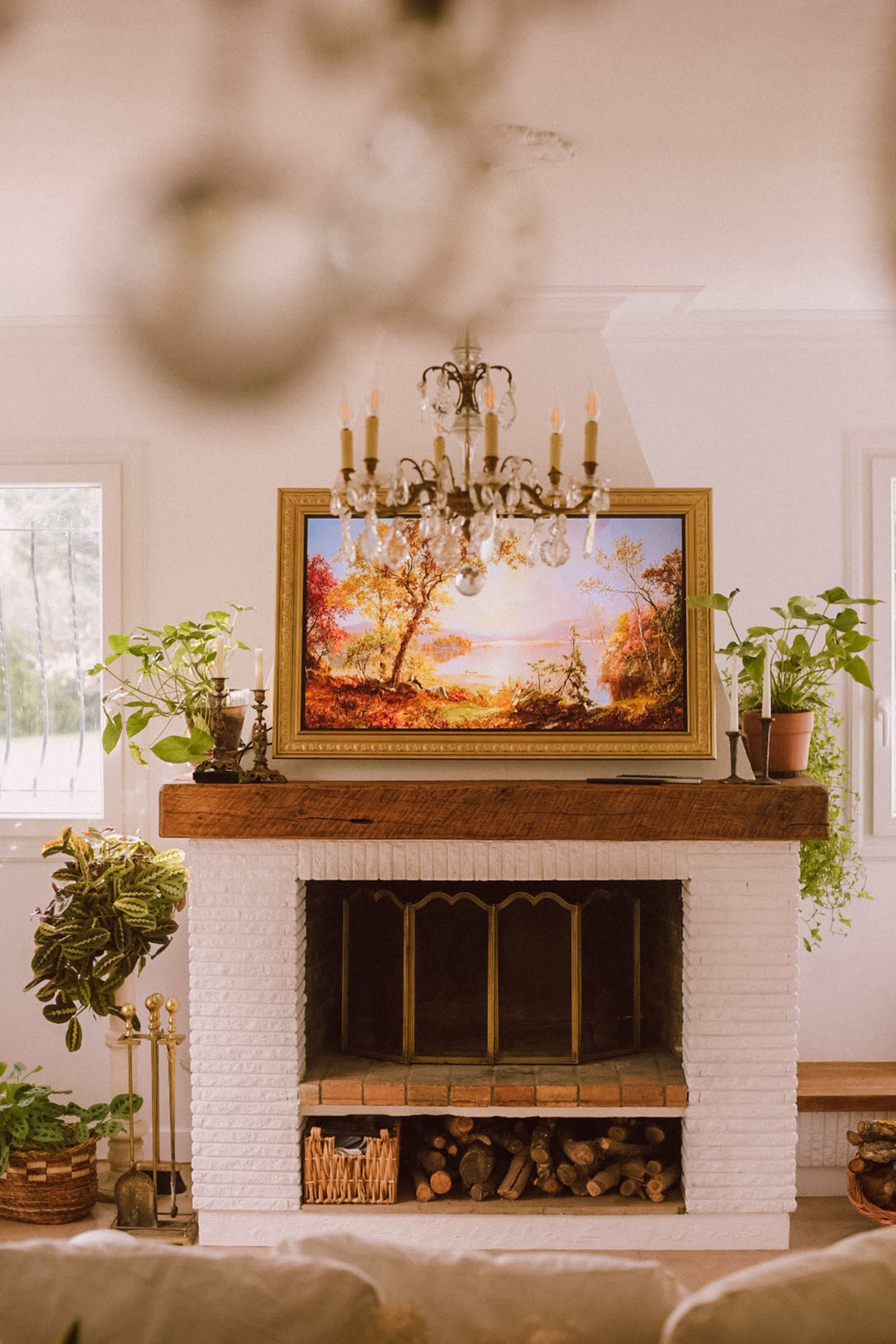 A chandelier hangs above a mantel adorned with a landscape painting, surrounded by potted plants and firewood.