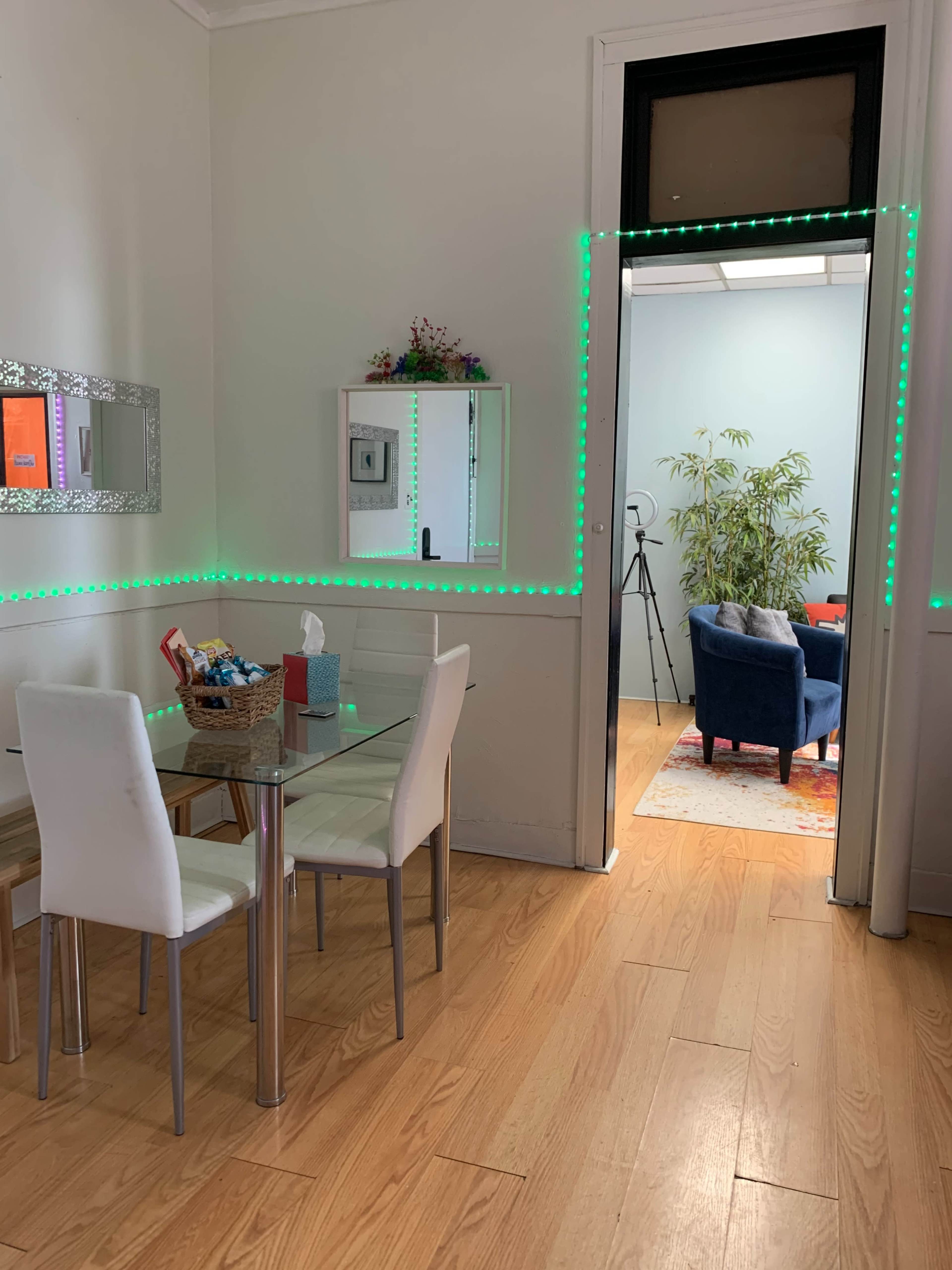A well-lit room with a glass dining table and white chairs, leading to another room featuring a blue couch and a bamboo plant.
