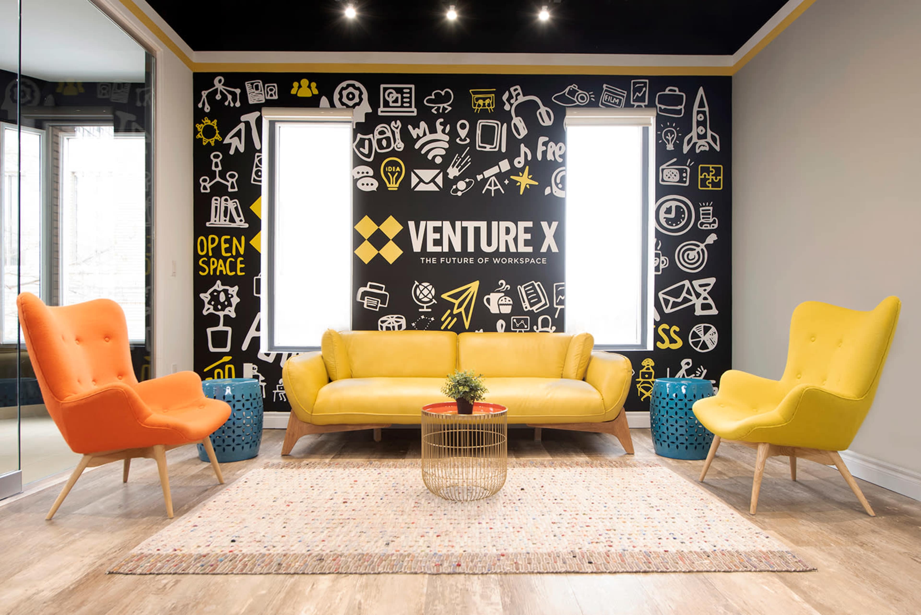 A modern workspace featuring a yellow couch, orange and yellow armchairs, a round coffee table, and a wall adorned with various black and white icons related to business and innovation.