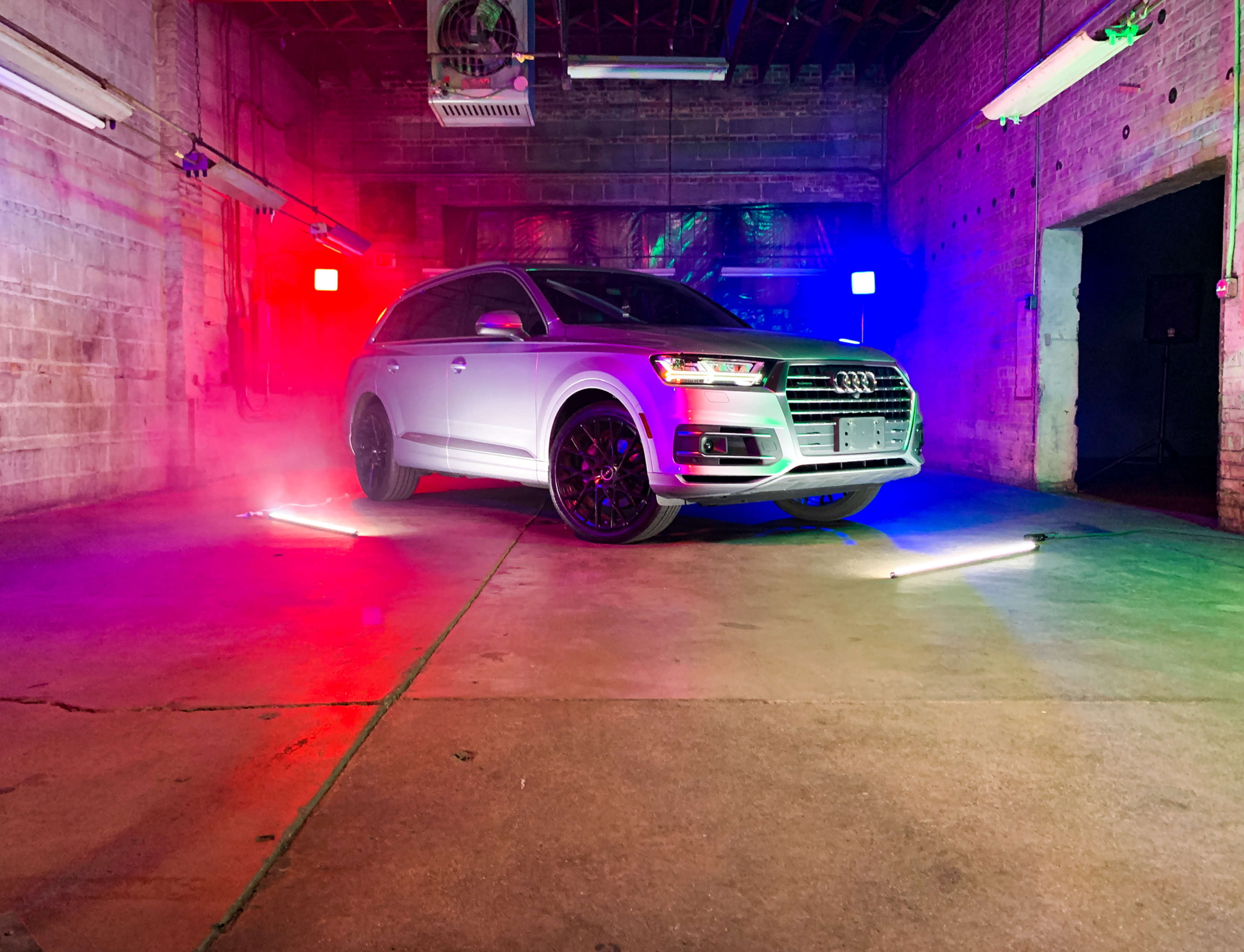 A silver Audi Q7 is positioned in a dimly lit industrial space, illuminated by red, blue, and green lights.