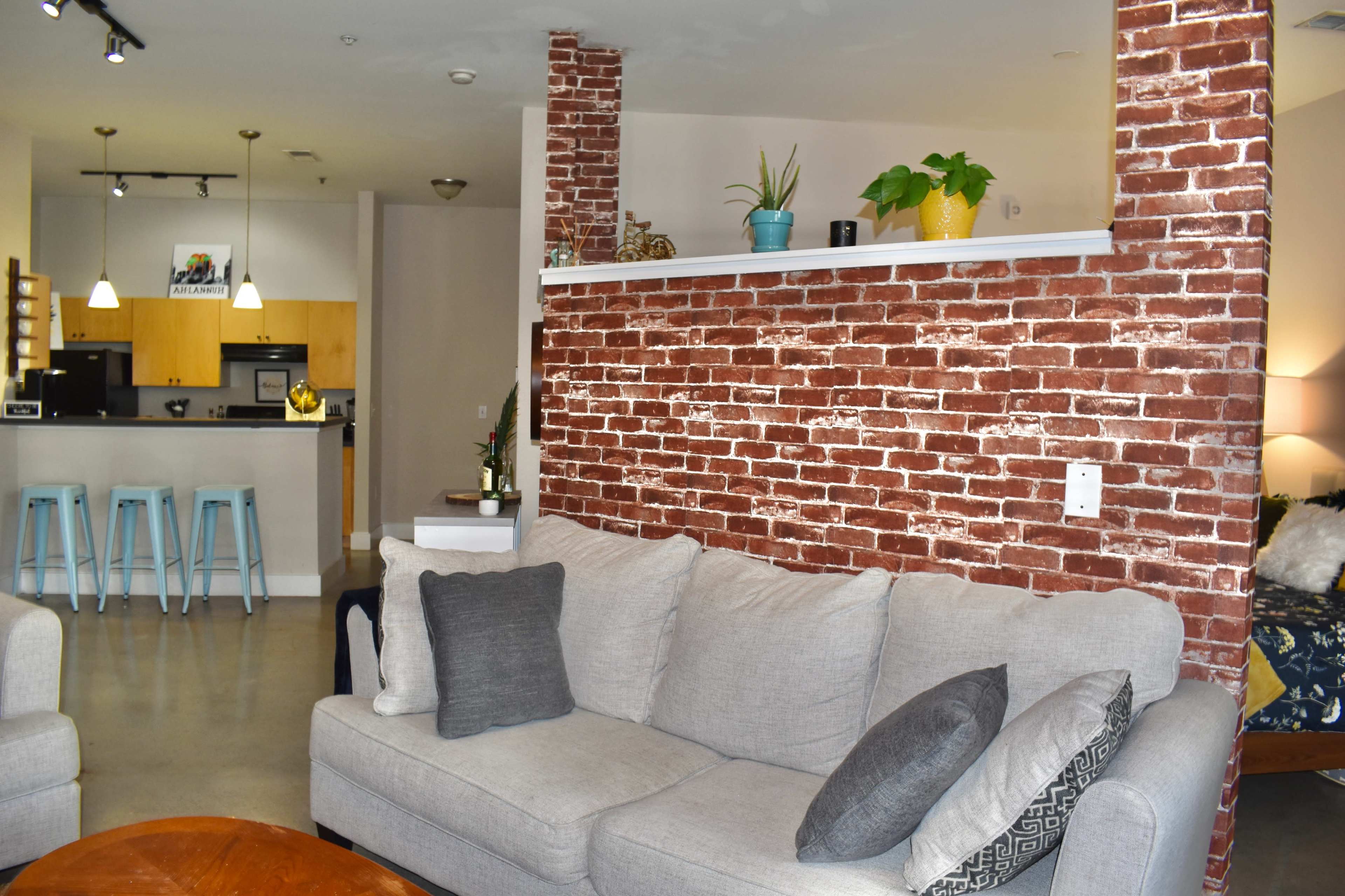 A modern living room with a brick accent wall behind a grey sofa, a wooden coffee table in the foreground, and a kitchenette visible in the background.