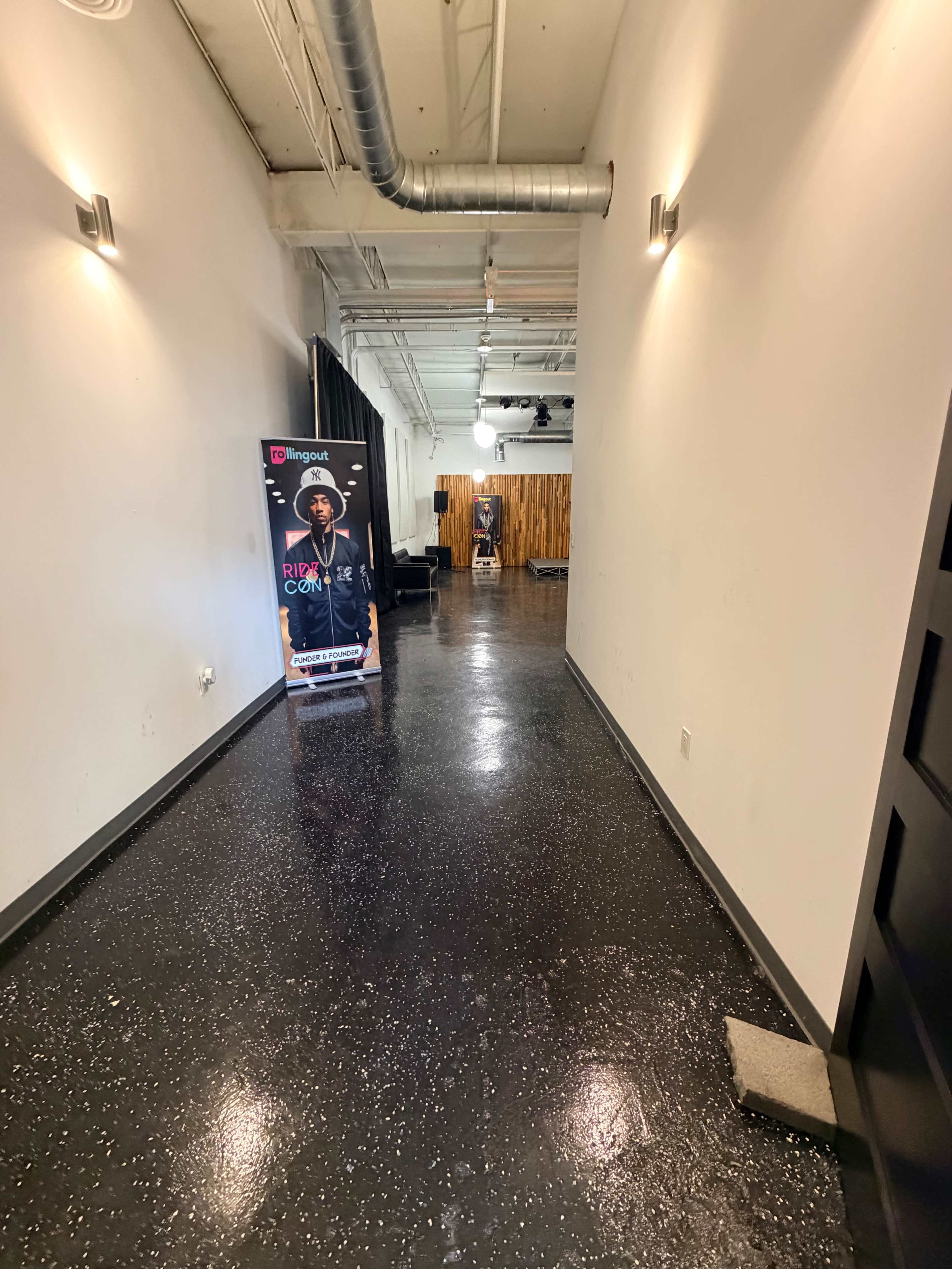 The image shows a long, narrow hallway with polished black floors, lighting fixtures on the walls, and a promotional stand featuring a performer in the foreground.