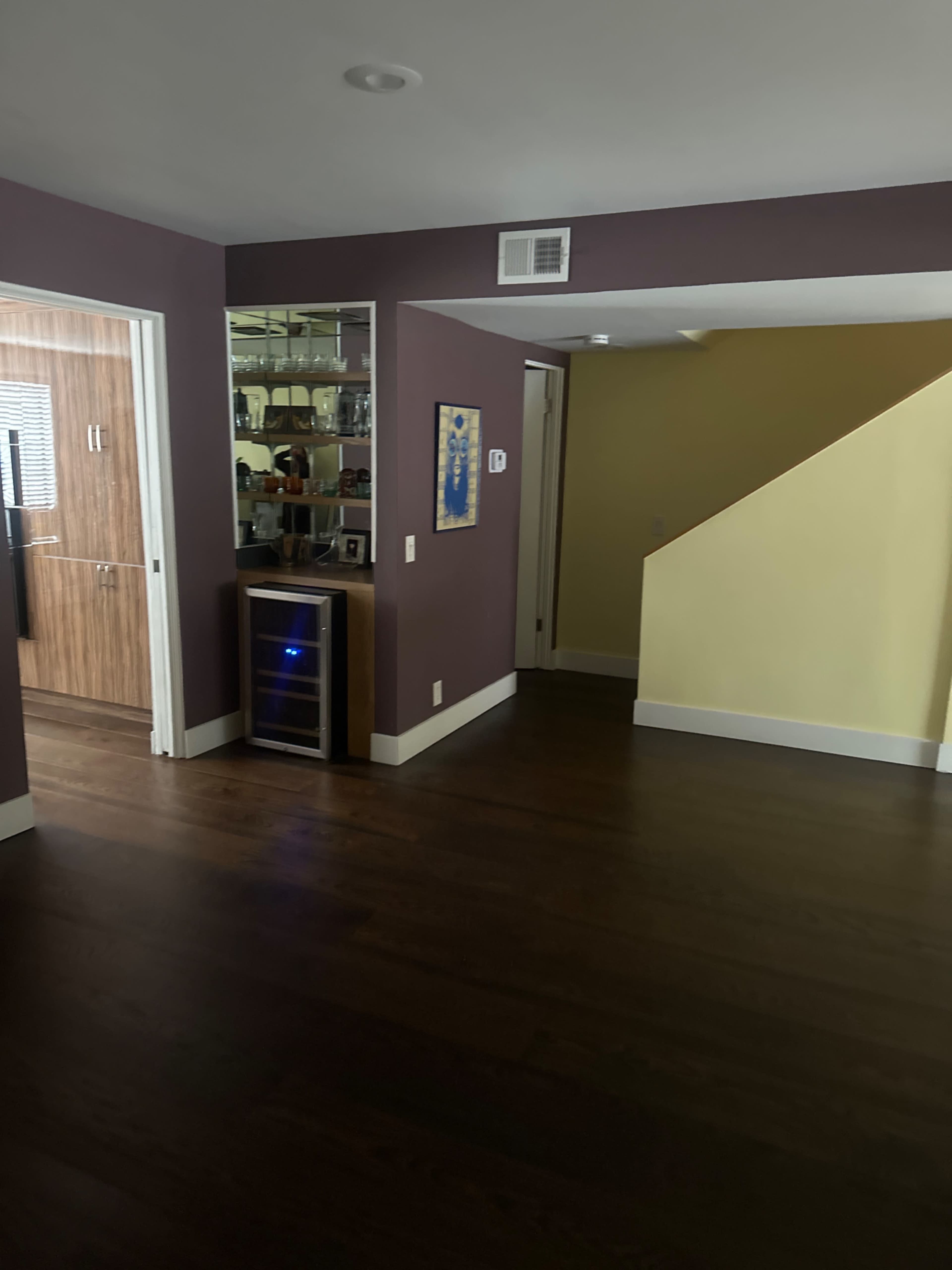 The image shows a well-lit interior space featuring a purple wall, a wine cooler against one wall, a staircase, and an open doorway leading to another room.