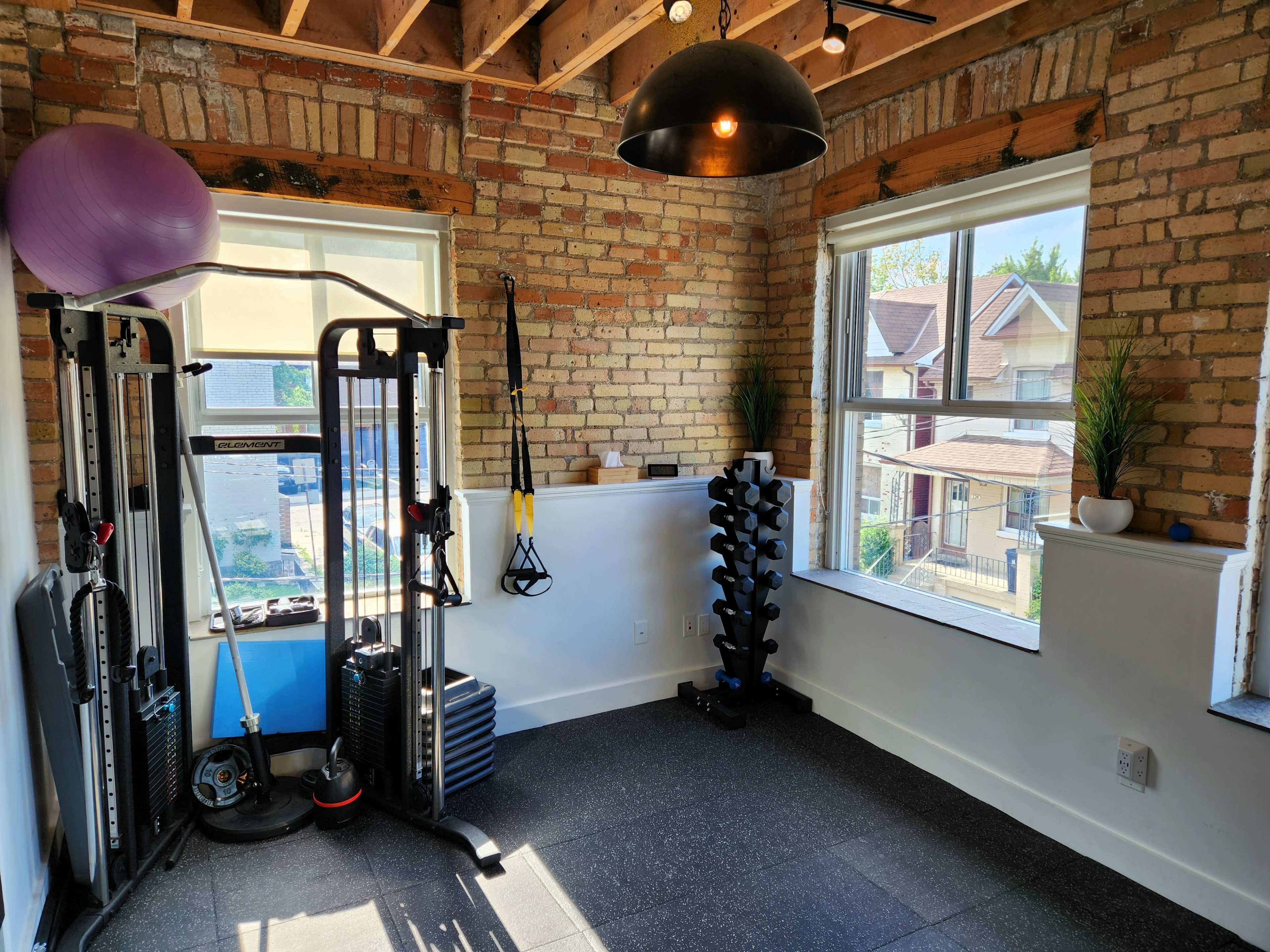 A home gym featuring exercise equipment, including a weight rack, a suspension trainer, and a stability ball, with large windows and exposed brick walls.