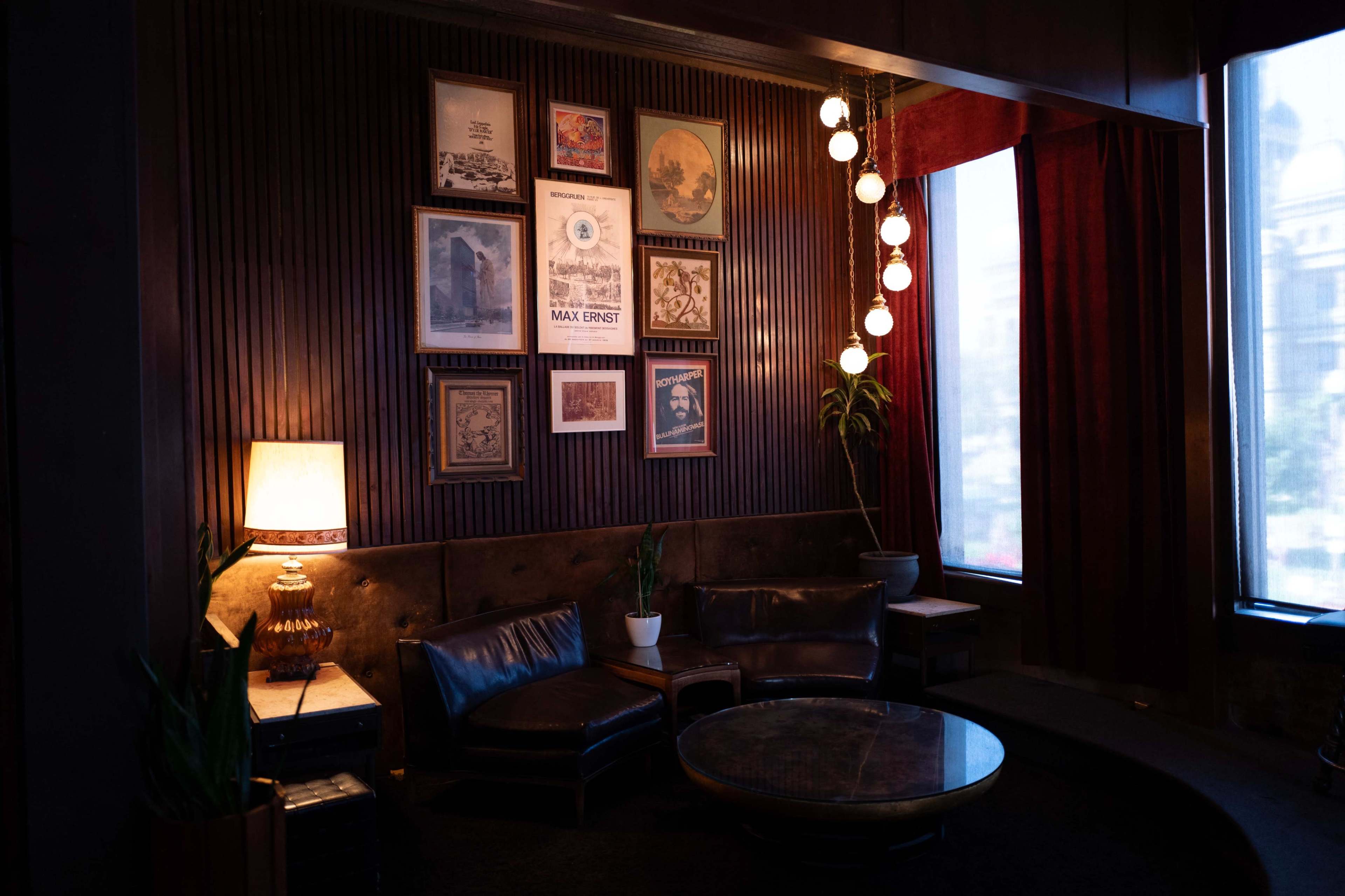 The image features a cozy lounge area with a dark wooden wall, framed artwork, a lamp, and a circular coffee table surrounded by leather seating.