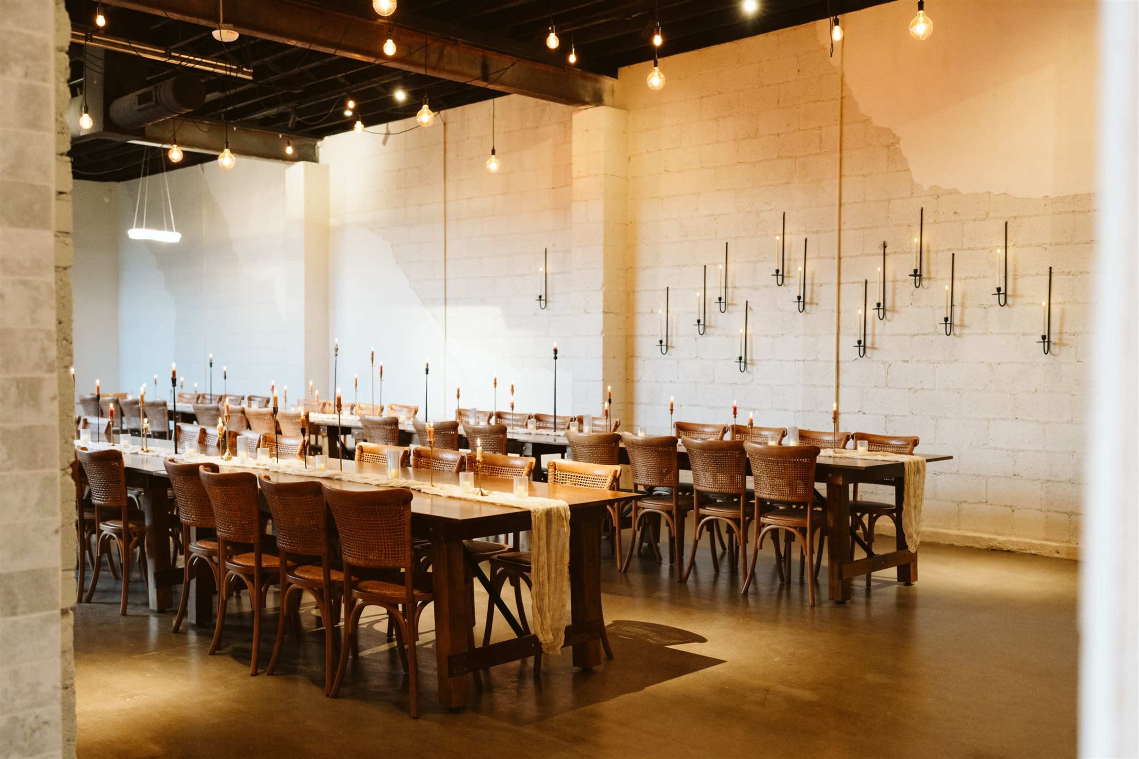 A spacious dining area features several long wooden tables arranged with chairs, illuminated by hanging light bulbs and wall-mounted candle holders.