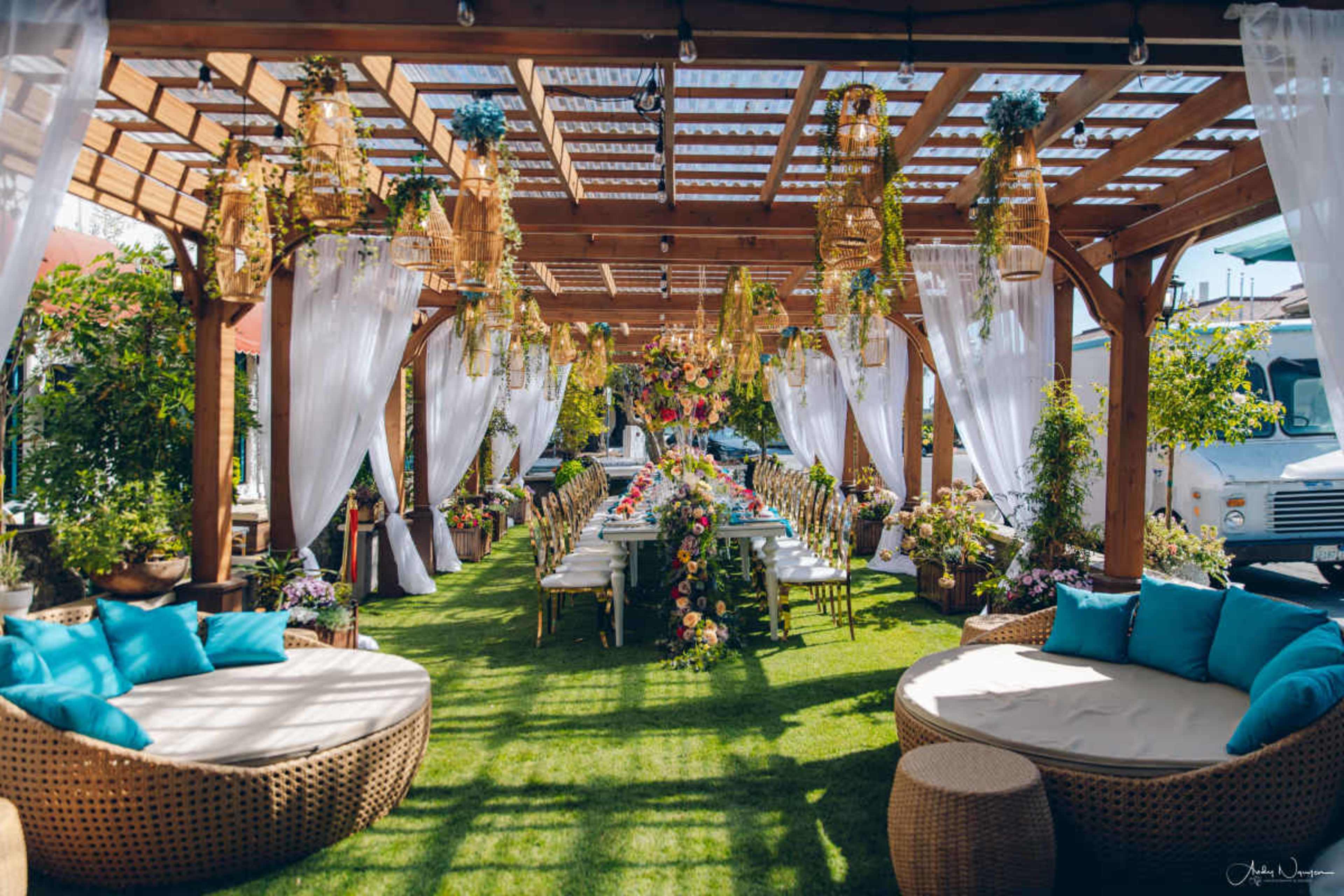 A beautifully decorated outdoor dining area features a long table set for a gathering, surrounded by lush greenery and vibrant floral arrangements under a wooden pergola.