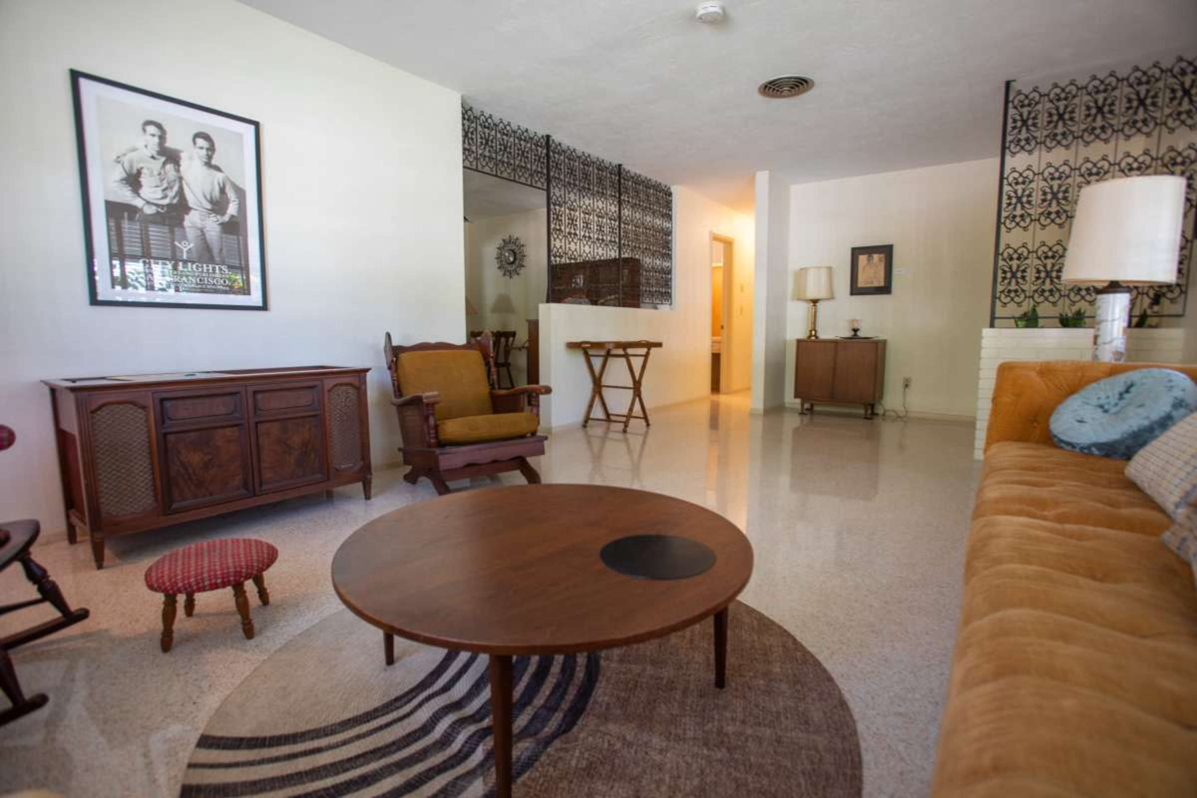 The image shows a mid-century modern living room with a beige sofa, a round coffee table on a patterned rug, and a wooden sideboard, all set in a bright space with light-colored walls.