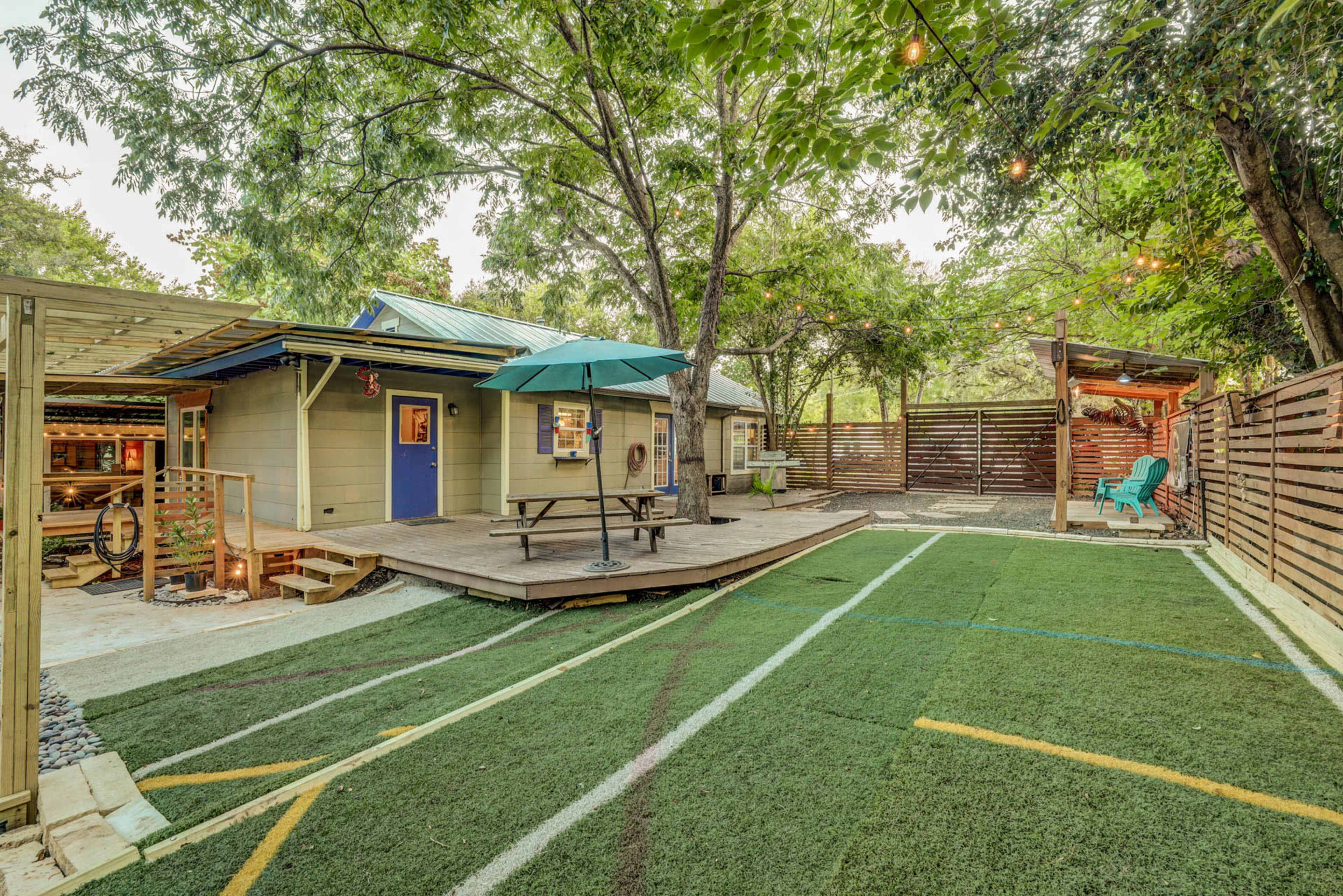 A landscaped backyard with artificial turf, a small deck with a table and umbrella, and a cozy seating area surrounded by wooden fencing and trees.