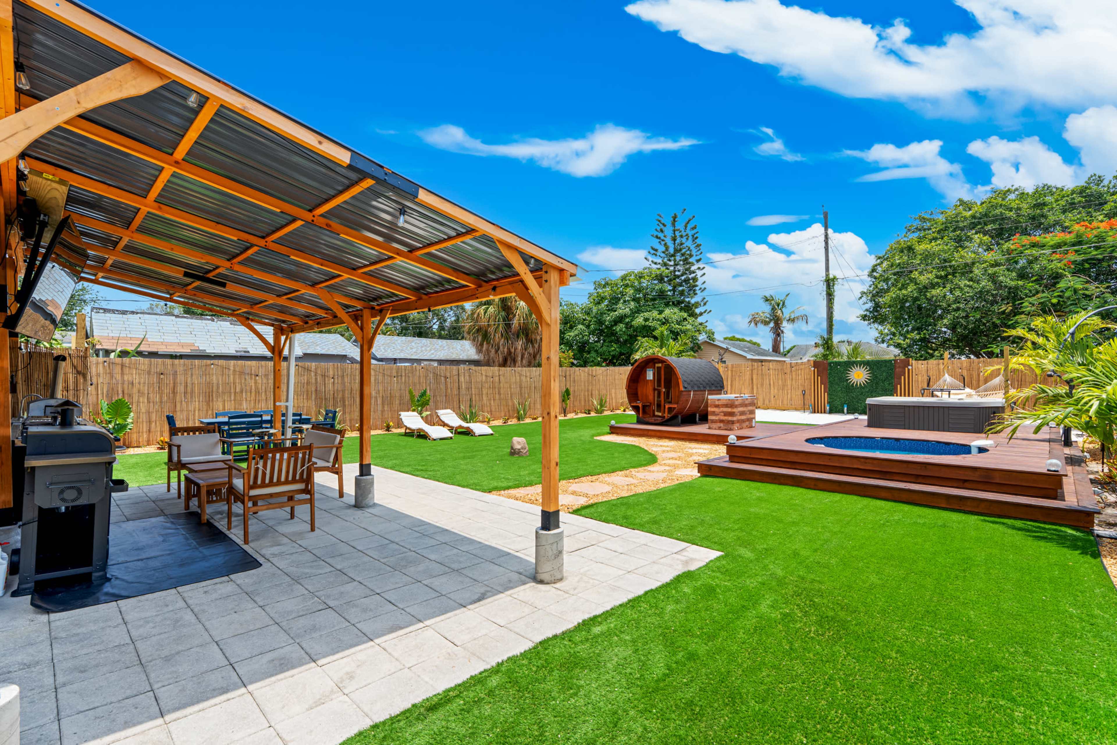 The image shows a well-maintained backyard featuring a covered seating area, a hot tub, a barrel sauna, and manicured artificial grass.