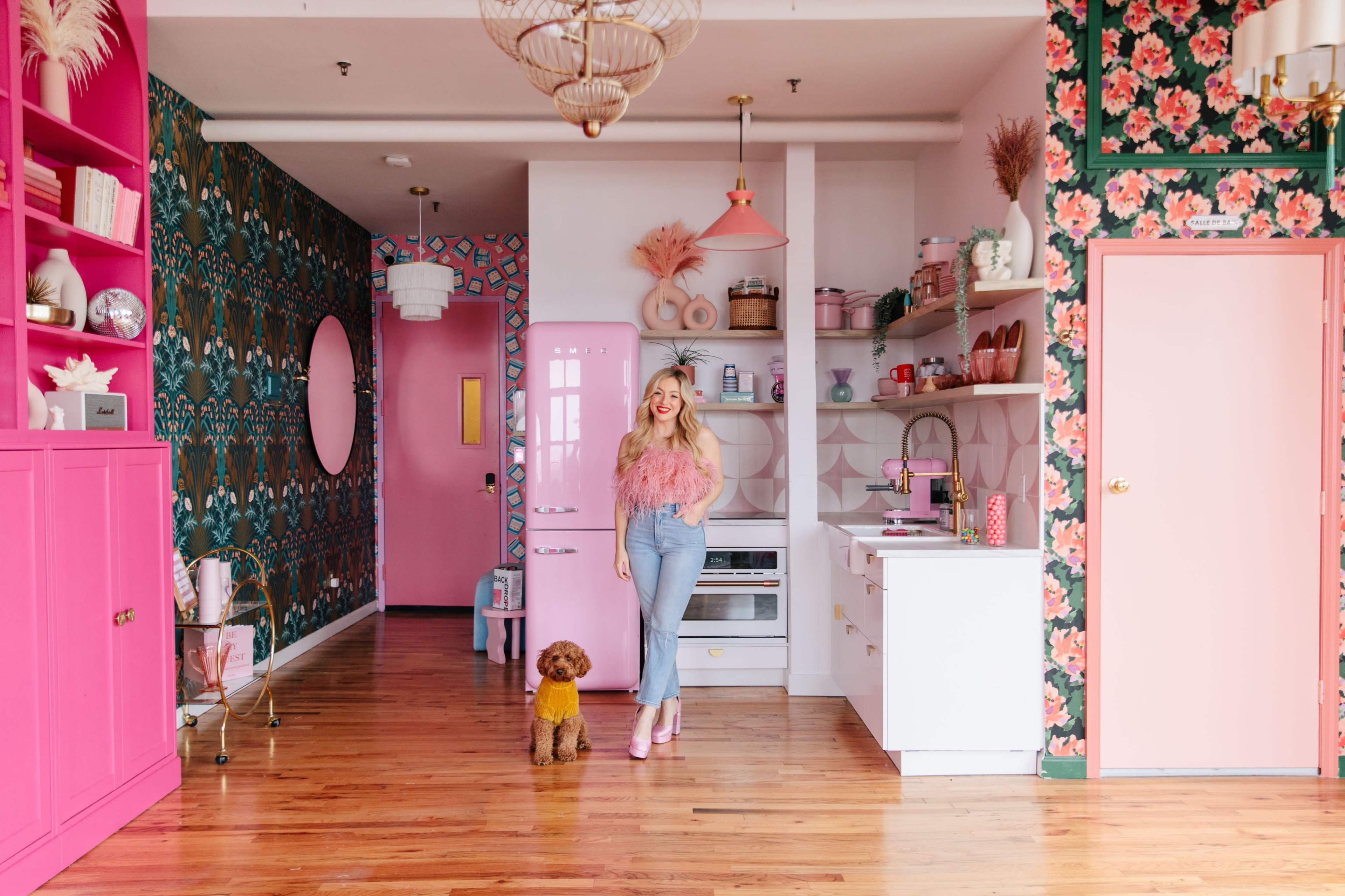 A woman stands in a brightly decorated kitchen with pink accents and floral wallpaper, next to a small brown dog.
