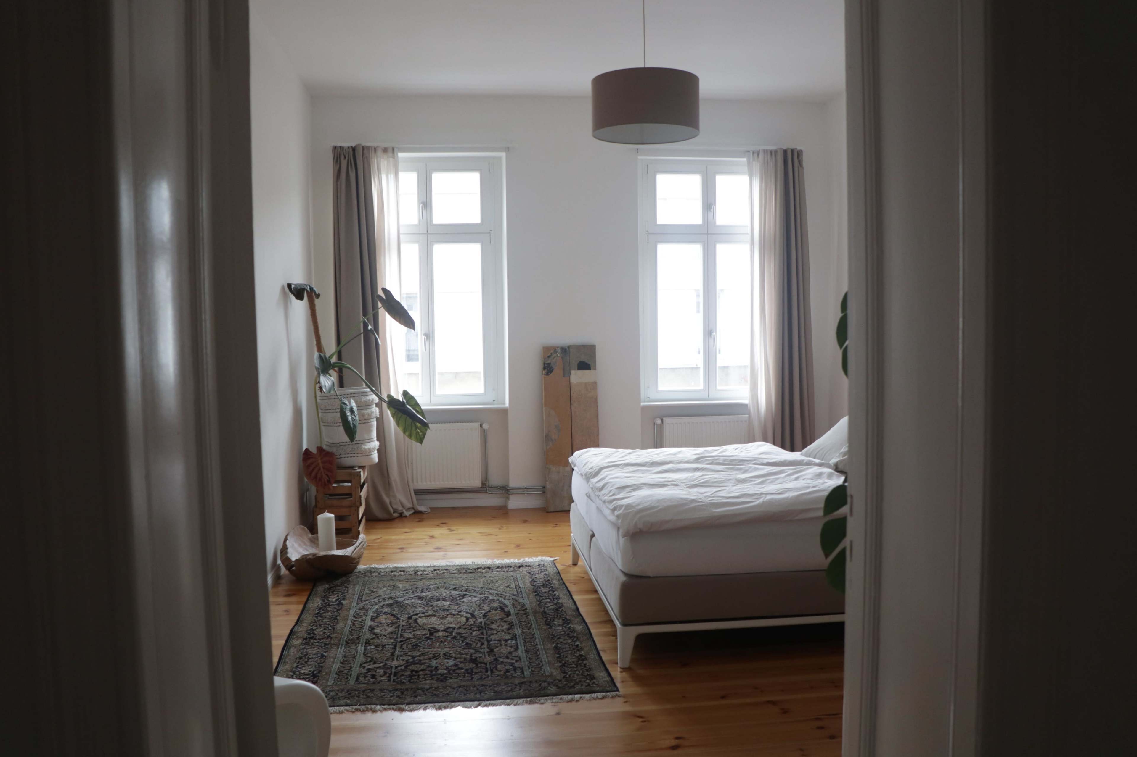 A bright bedroom features two windows, a bed with white bedding, a patterned rug, and indoor plants.