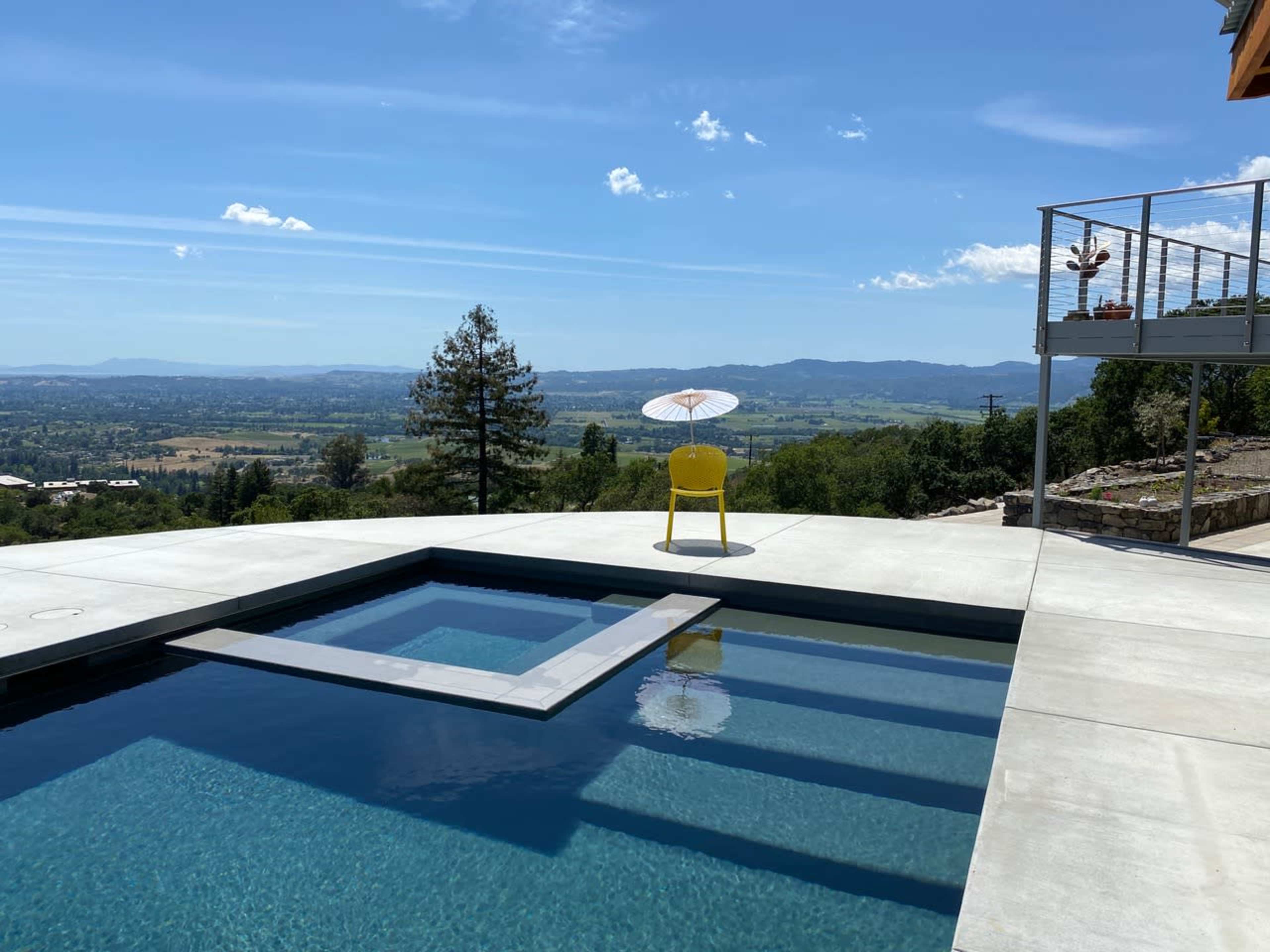 A yellow chair with a parasol sits next to a rectangular swimming pool overlooking a scenic valley.