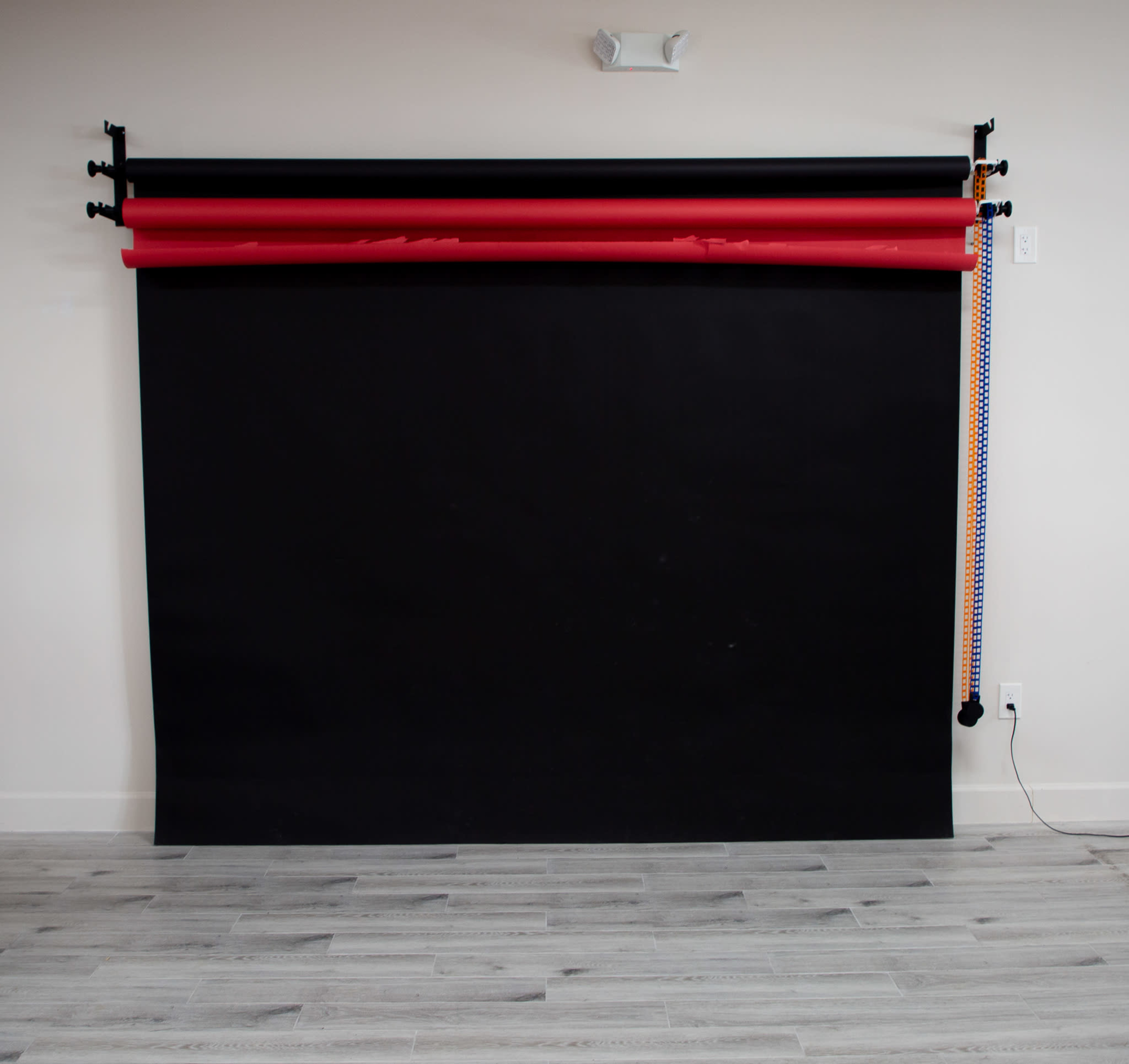 A dark backdrop setup with red and orange studio rolls suspended from a bar on a wall, with wooden flooring visible below.