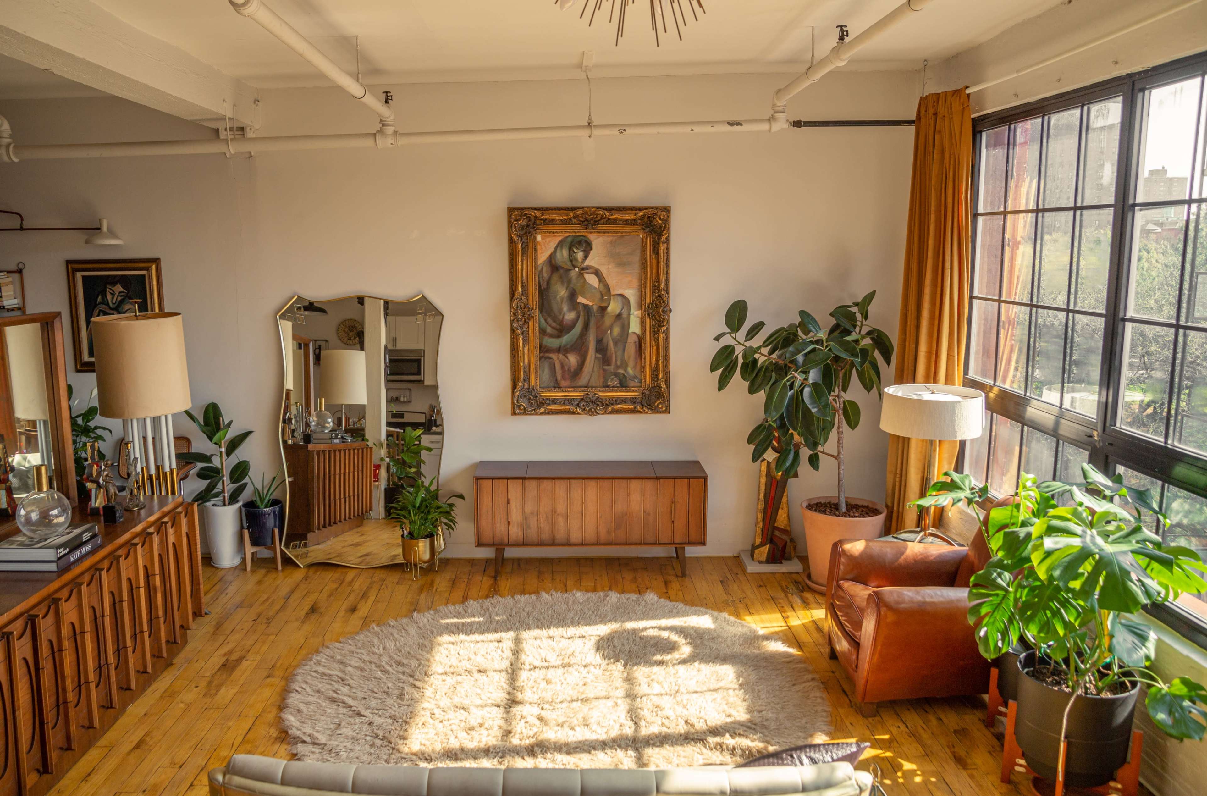 A bright living room features a large painting on the wall, a round rug on wooden flooring, potted plants, and mid-century furniture including a leather armchair and a wooden sideboard.