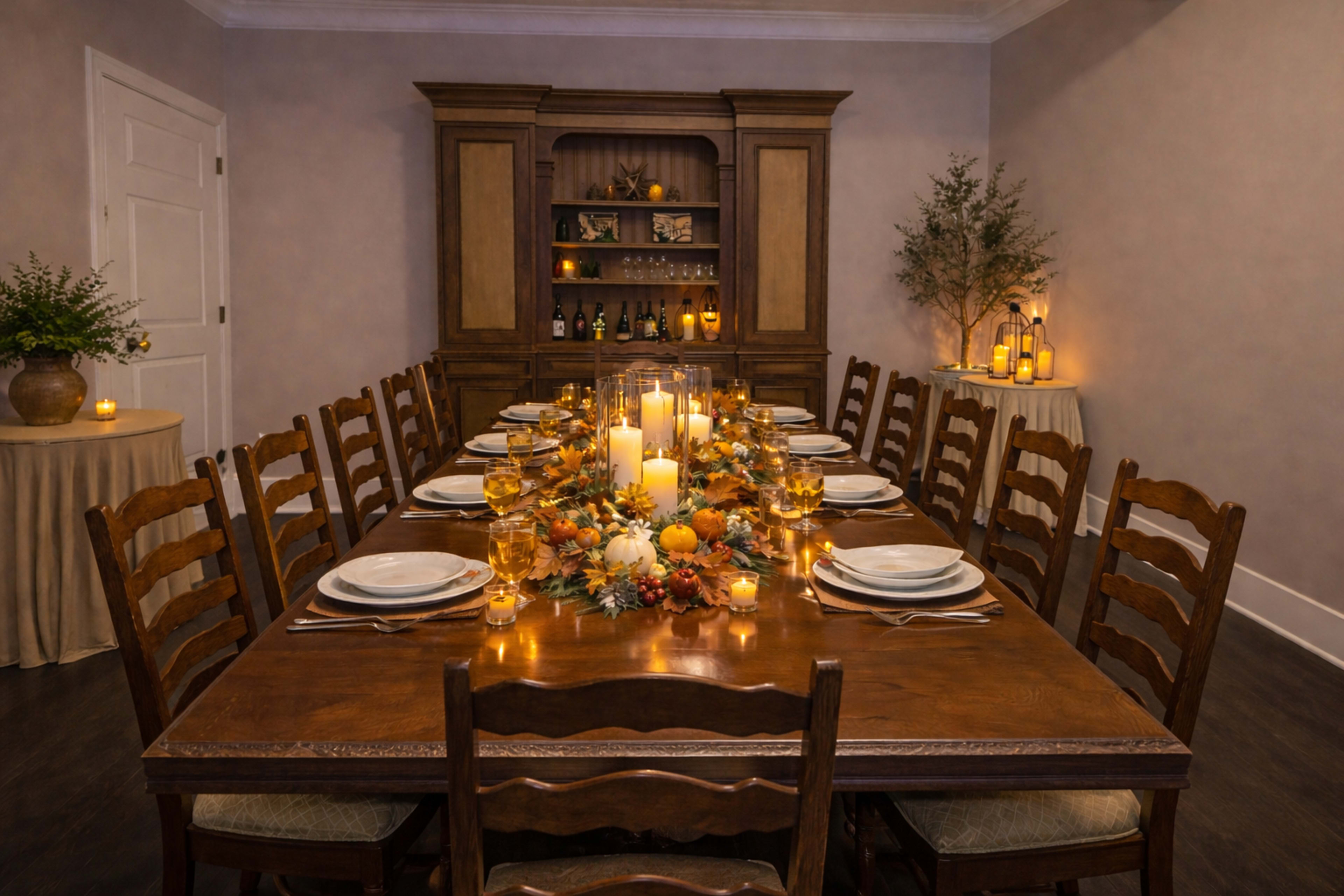 A long wooden dining table is set with plates and glasses, featuring a fall-themed centerpiece of candles and seasonal decorations, in a warmly lit dining room.