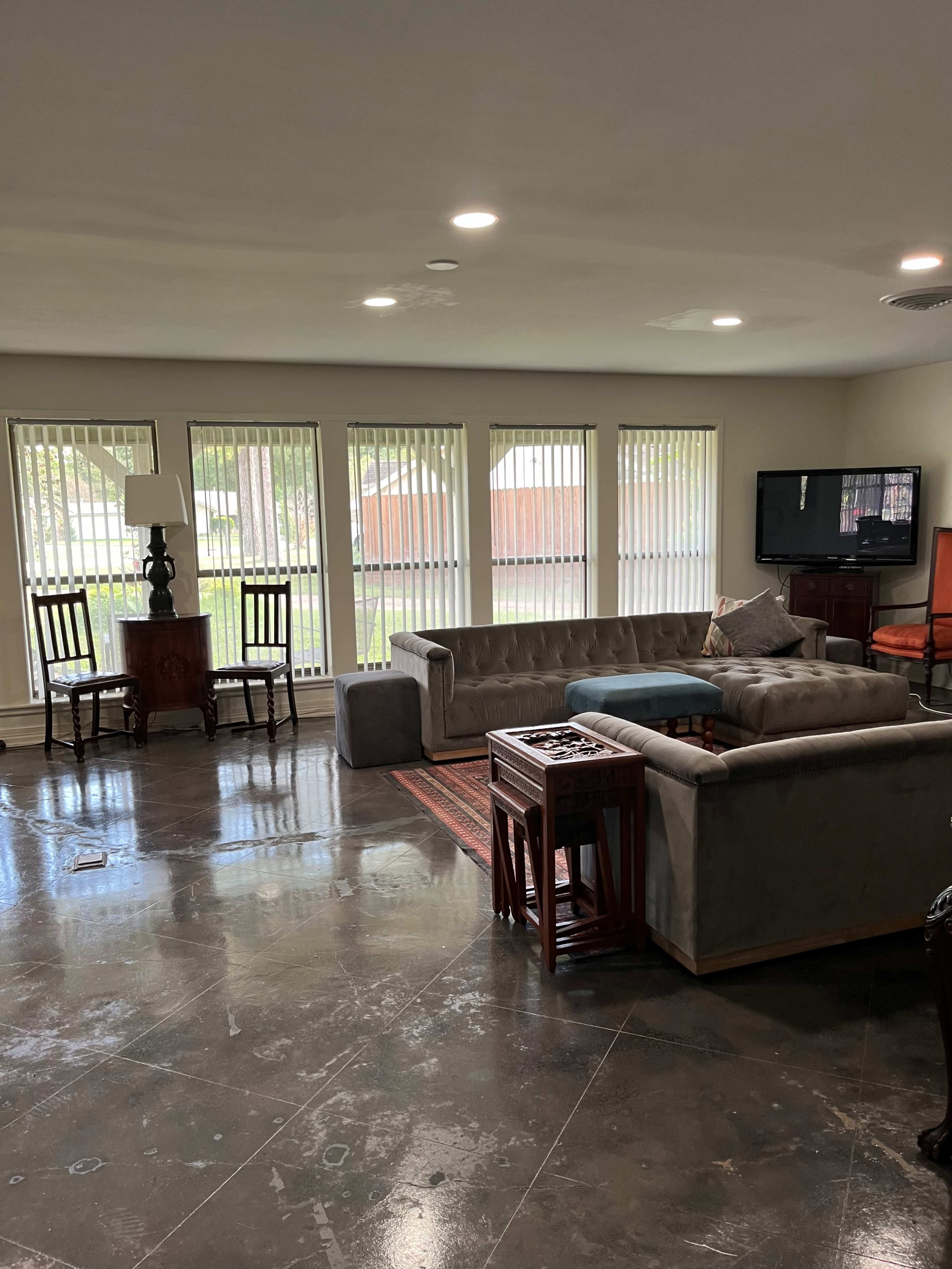 A spacious living room with large windows, a sectional sofa, two wooden chairs, a coffee table, and a television.