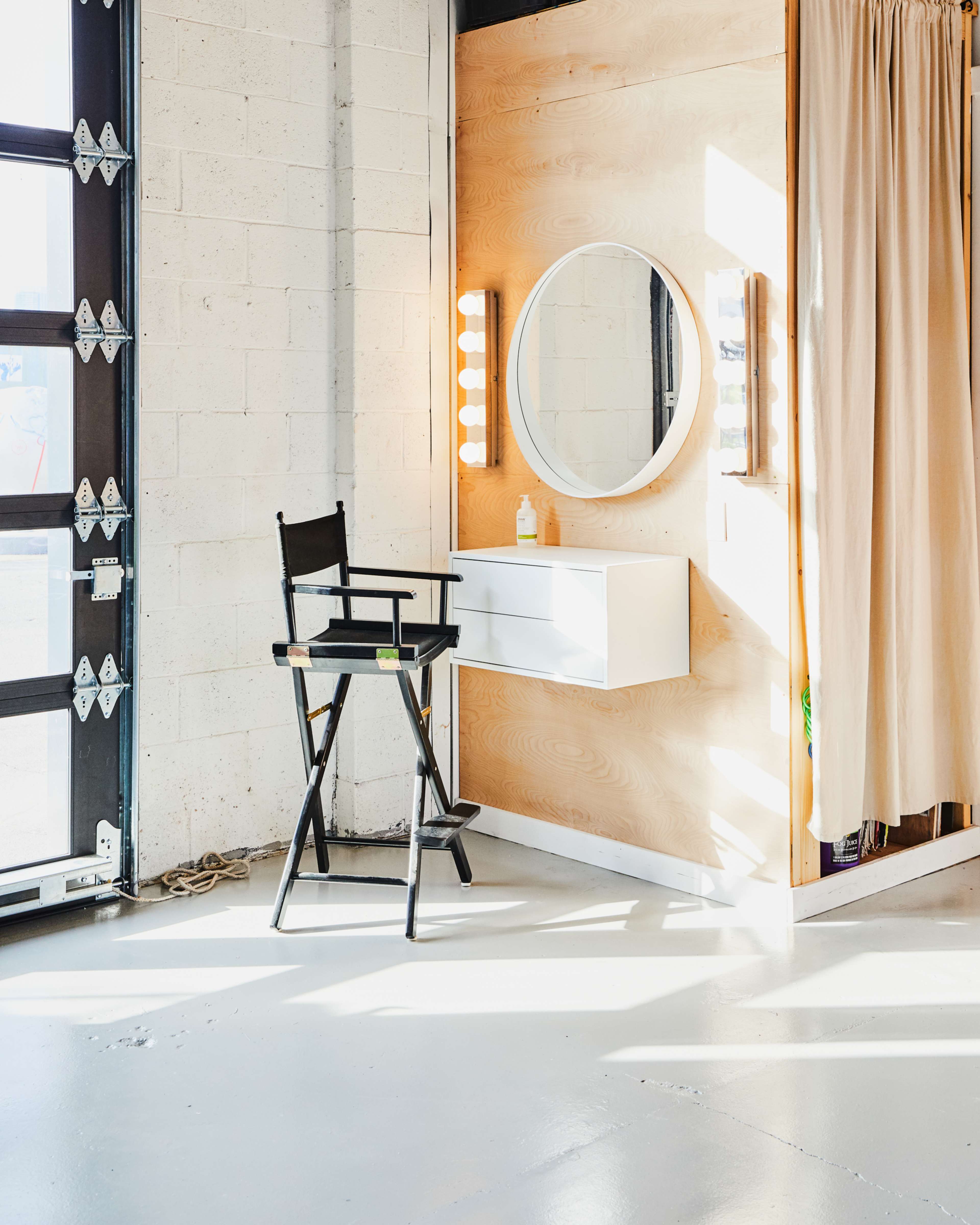 A well-lit makeup station with a circular mirror, a white shelf, and a director's chair against a wooden wall.