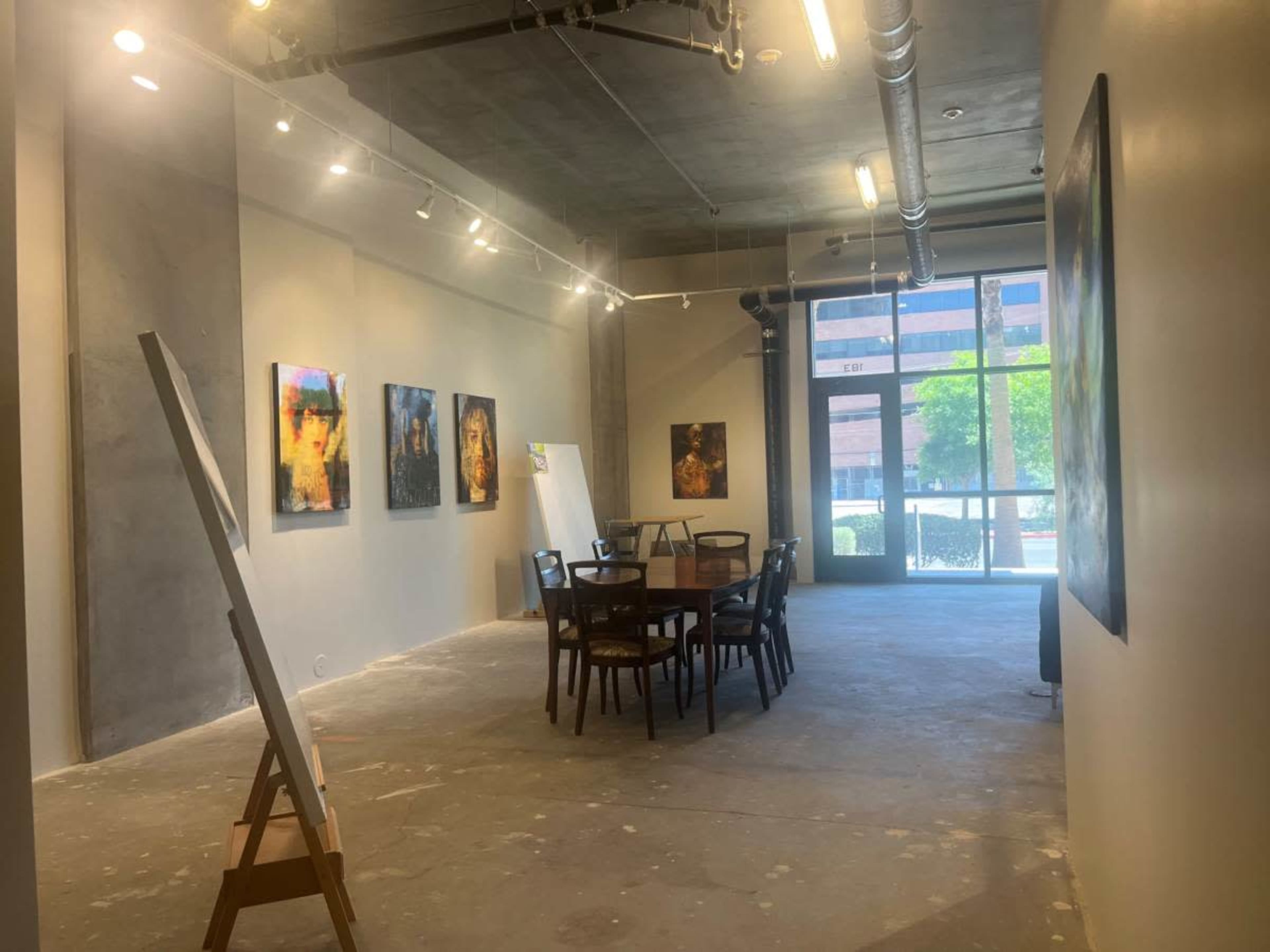 The image shows an art gallery featuring several paintings on the walls, a round table with chairs, and large windows letting in natural light.