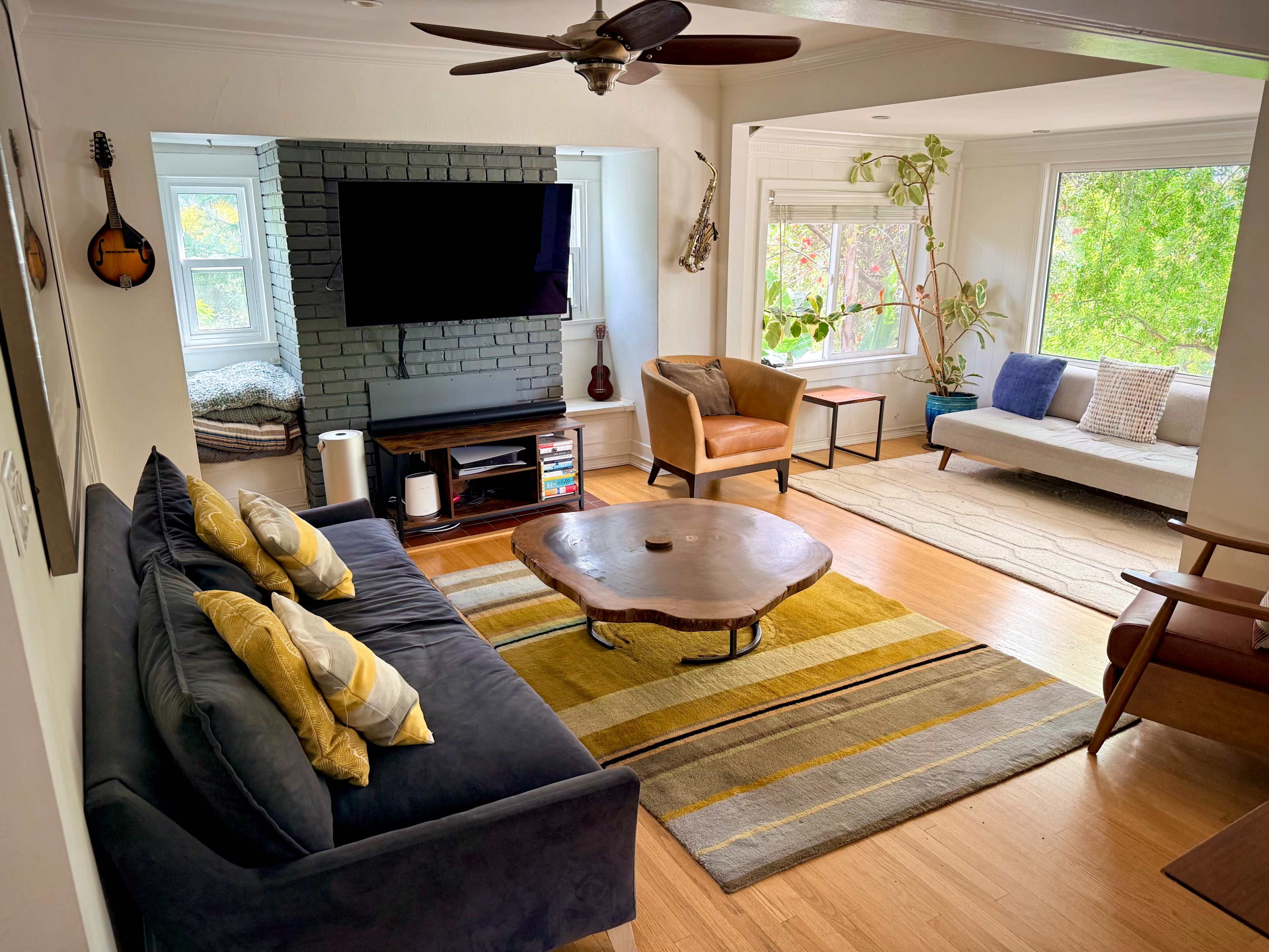 The image shows a living room with a dark sofa, a wooden coffee table, a television mounted on a brick wall, and a separate seating area with a light sofa and armchair near large windows.