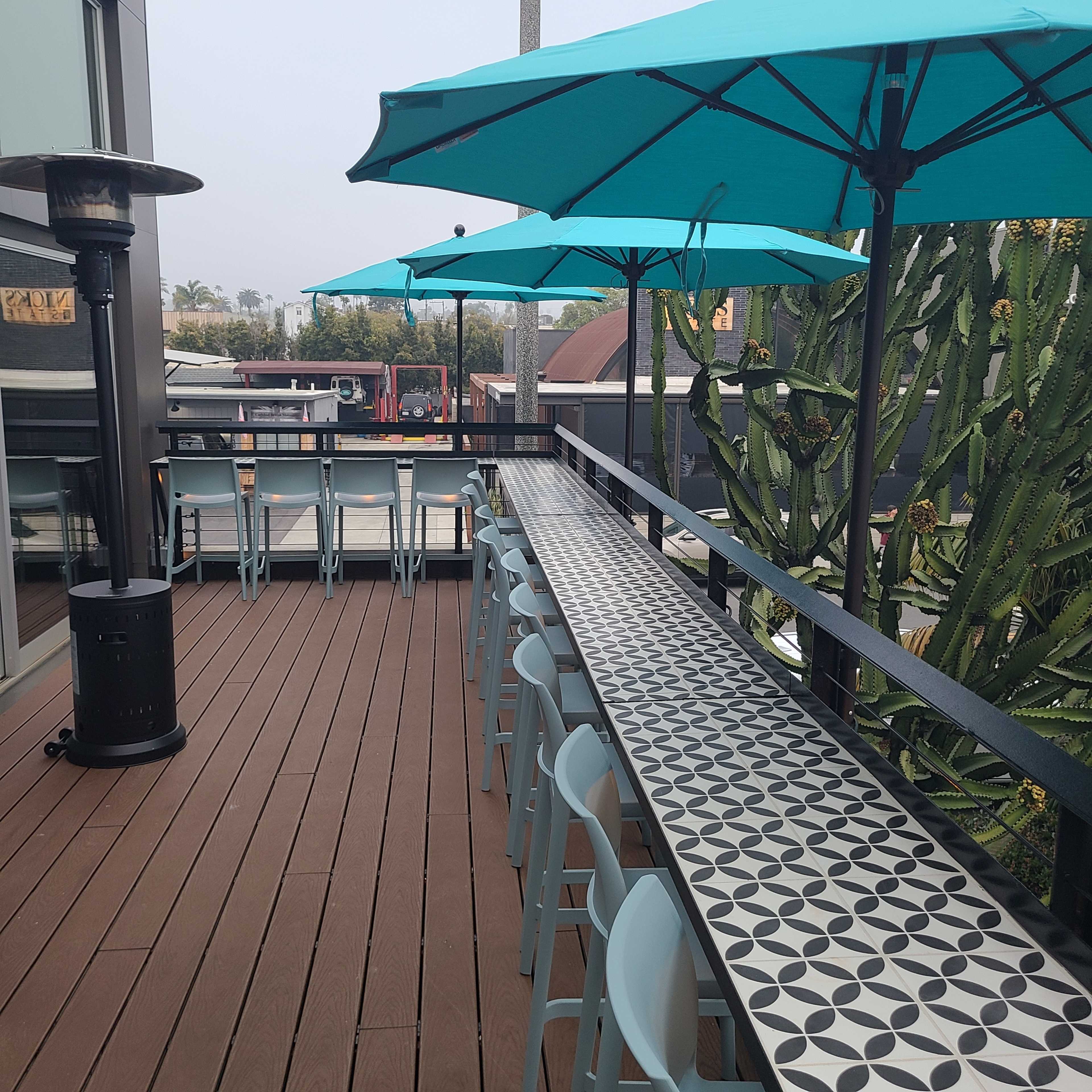 The image shows a rooftop patio with blue umbrellas, a long patterned countertop, and wooden flooring.