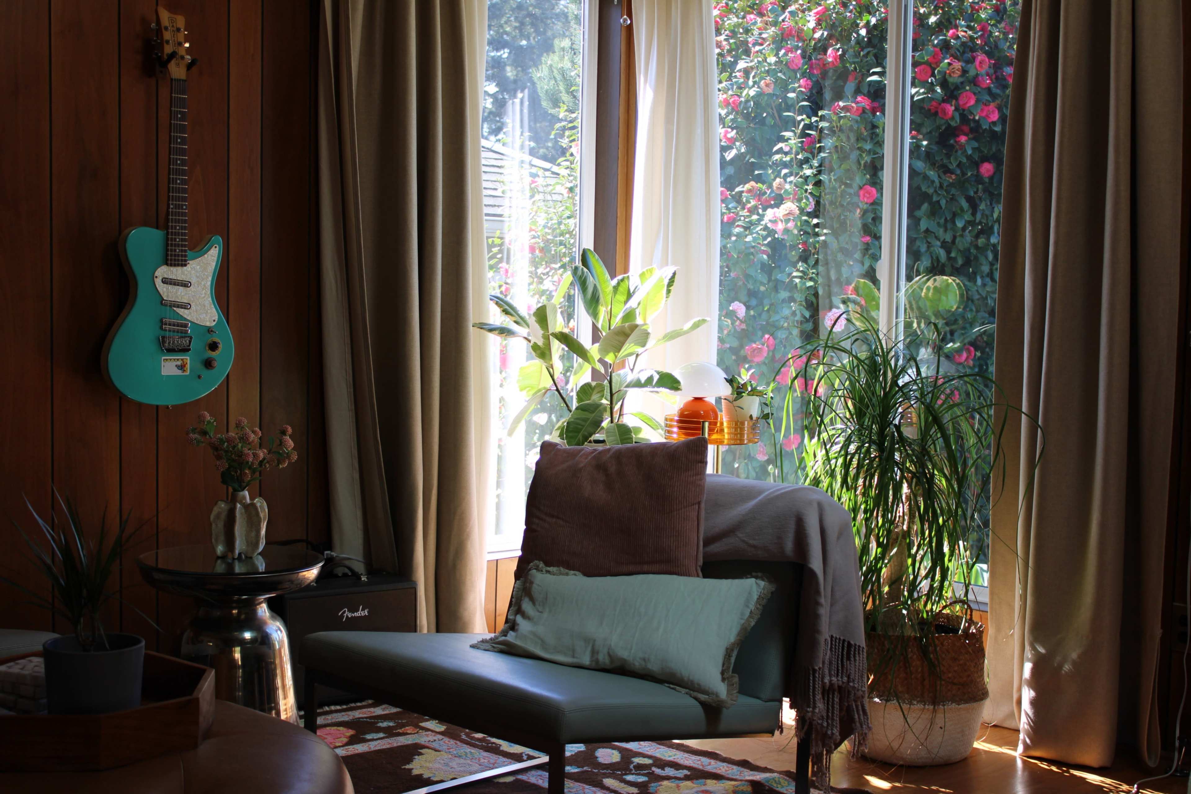A turquoise guitar hangs on a wooden wall beside a chair with cushions, while sunlight filters through large windows adorned with plants and a colorful garden outside.