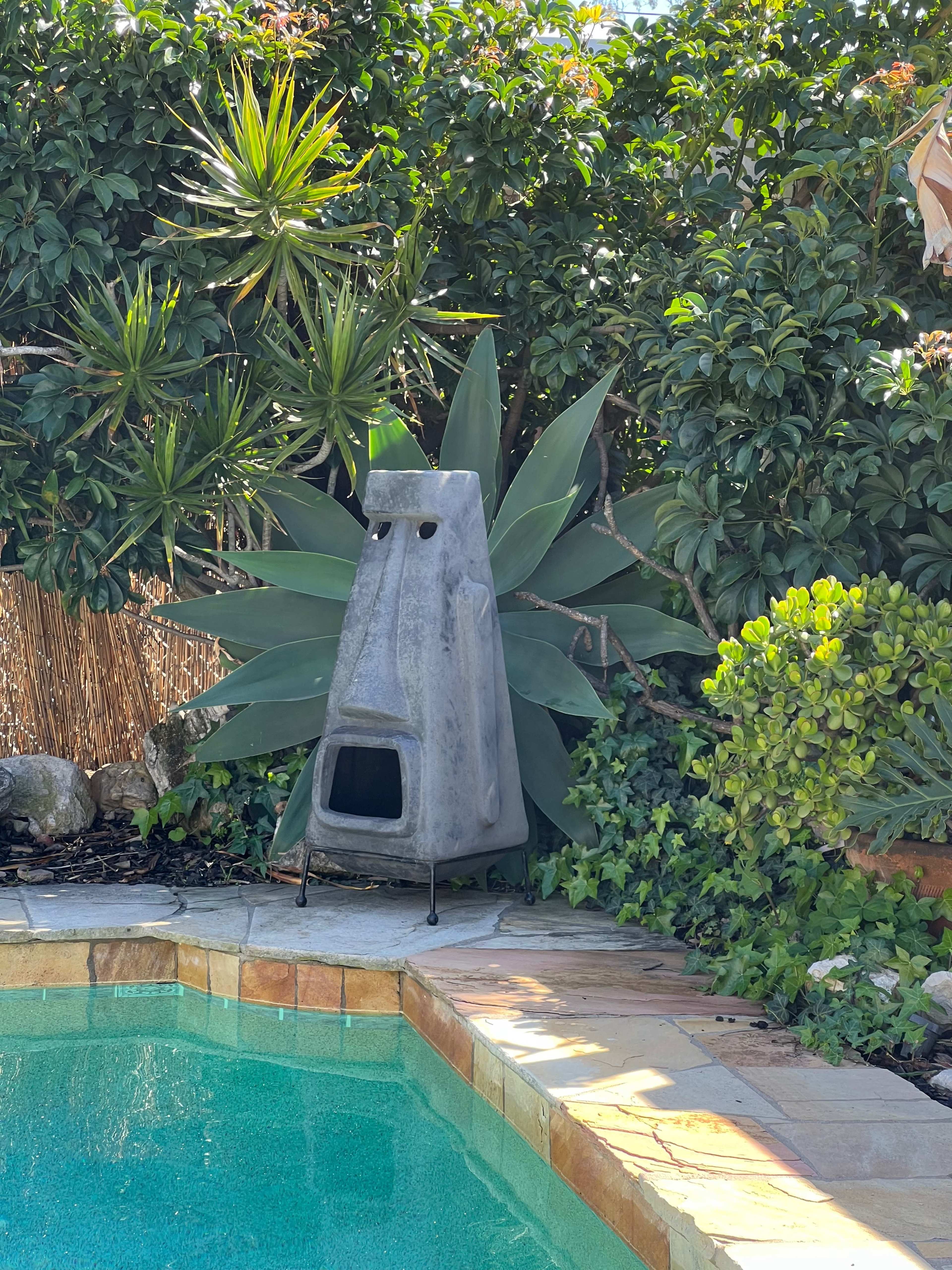 A concrete outdoor fireplace stands near a swimming pool, surrounded by lush greenery and plants.