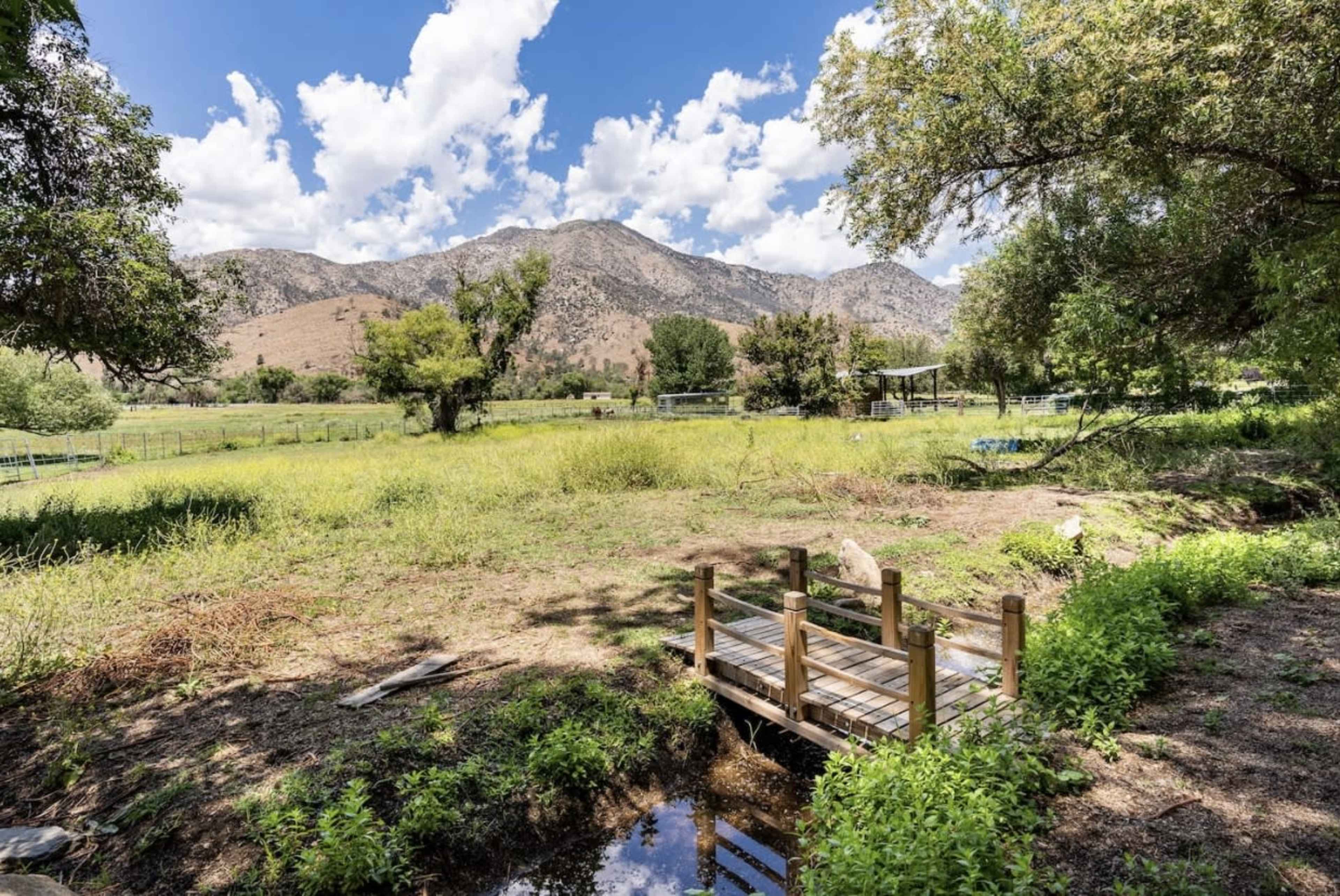 A wooden footbridge spans a shallow stream in a grassy area surrounded by mountains and scattered trees under a partly cloudy sky.