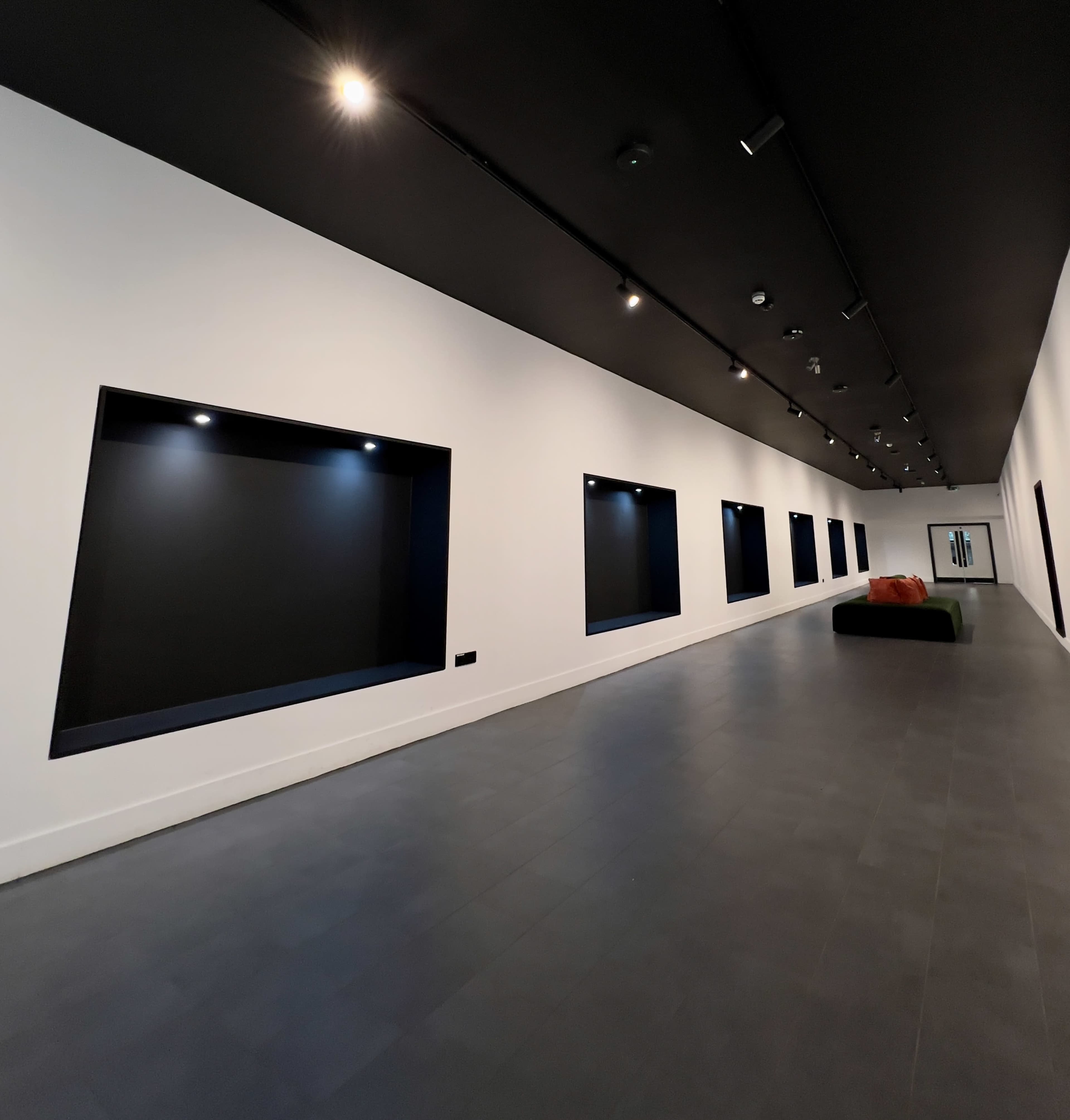 A minimalist gallery space with white walls, black recessed display areas, and a dark ceiling, illuminated by track lighting.