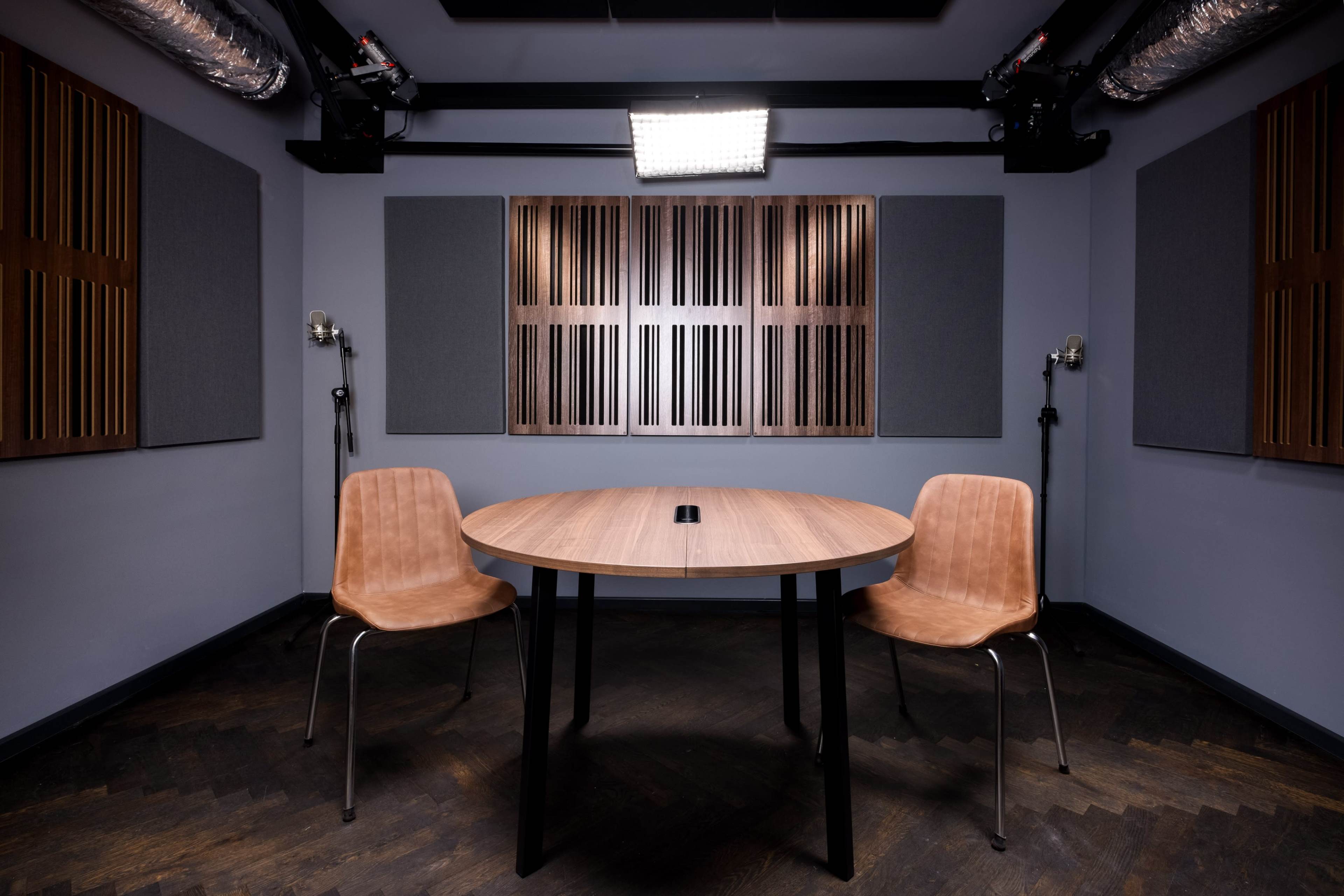 A small recording studio features a round wooden table at the center, flanked by two chairs, with soundproofing panels on the walls and ceiling lights above.